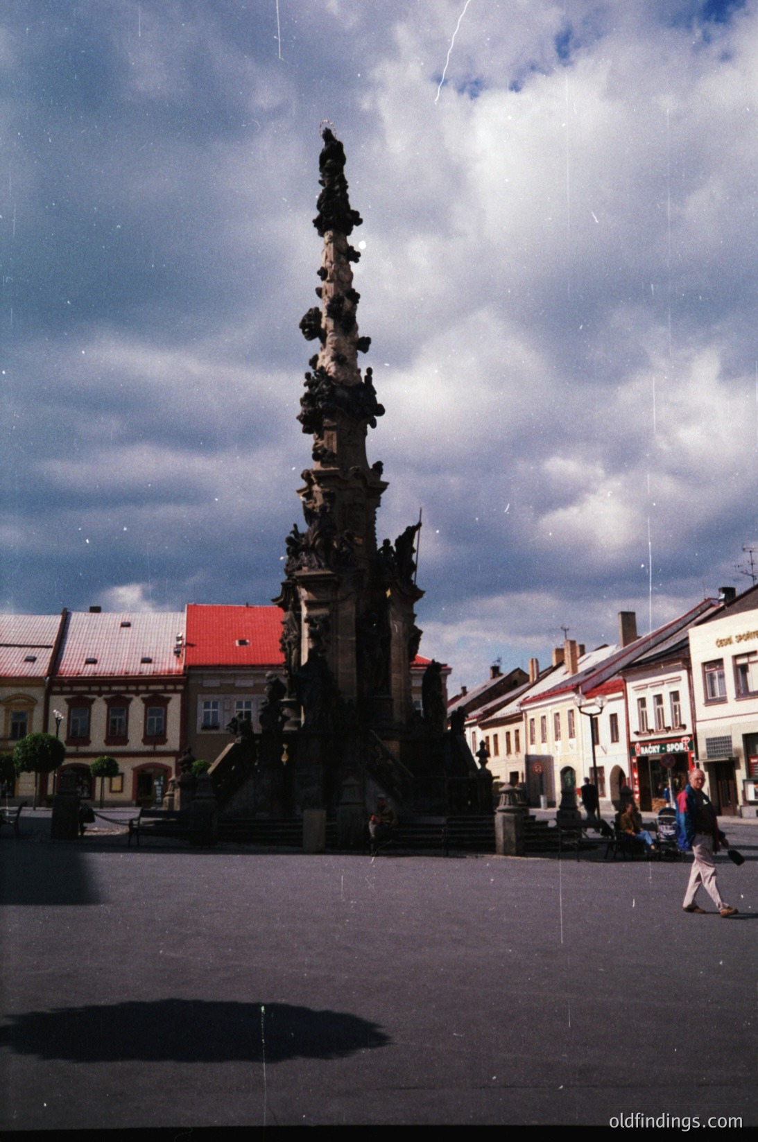 Baroque-style column monument with intricate sculptures, likely a 17th–18th century urban square. Surrounding buildings feature red-tiled roofs and classical facades. People casually stroll in the plaza, suggesting a public gathering space.