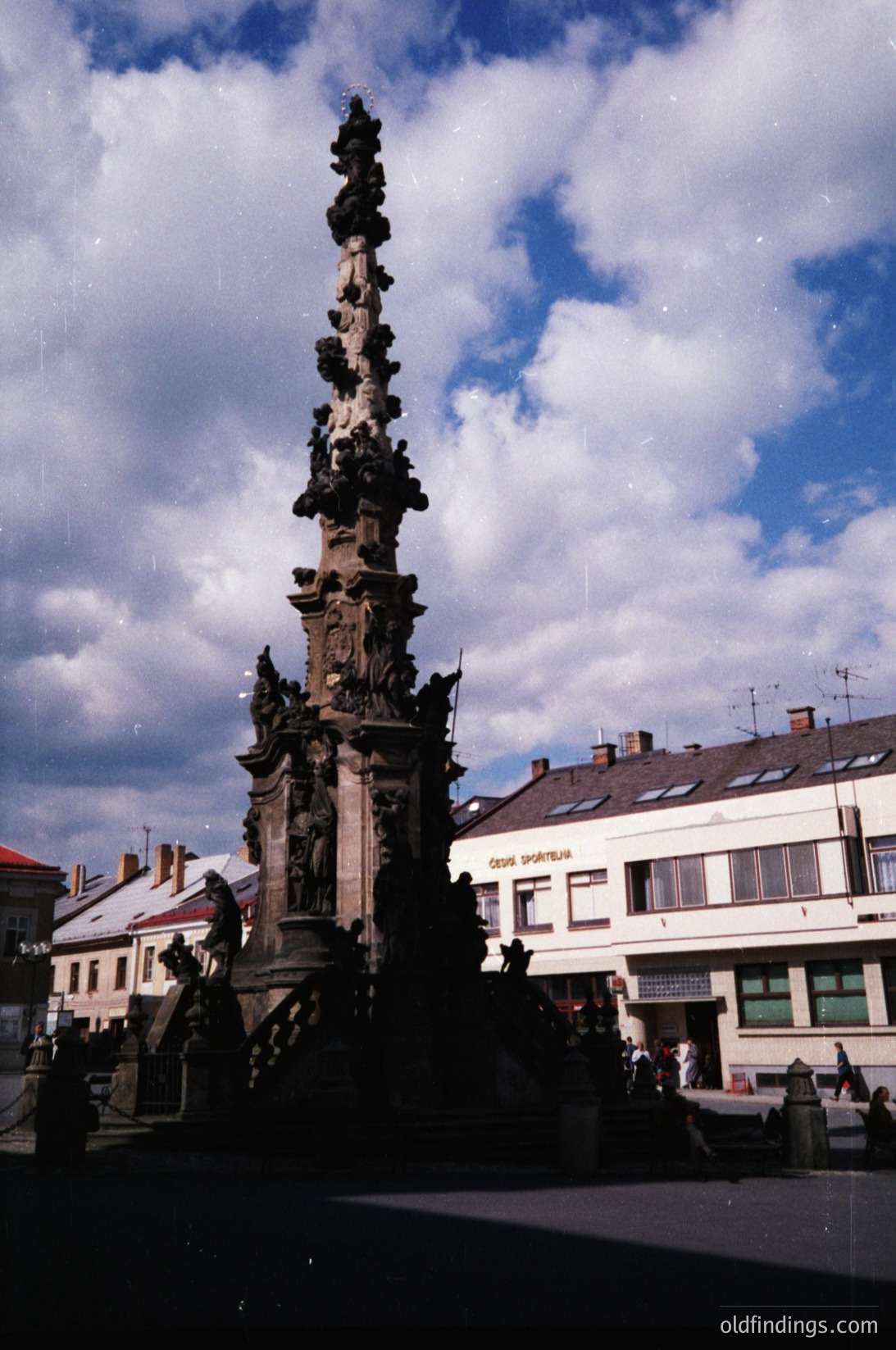 Baroque-style column monument with intricate carvings, likely a 17th–18th century urban fountain or memorial. Surrounding buildings feature classic European architecture with dormer windows and signage in Cyrillic script, suggesting Eastern Europe. Crowd of people gathered at base, indicating public square or tourist site.