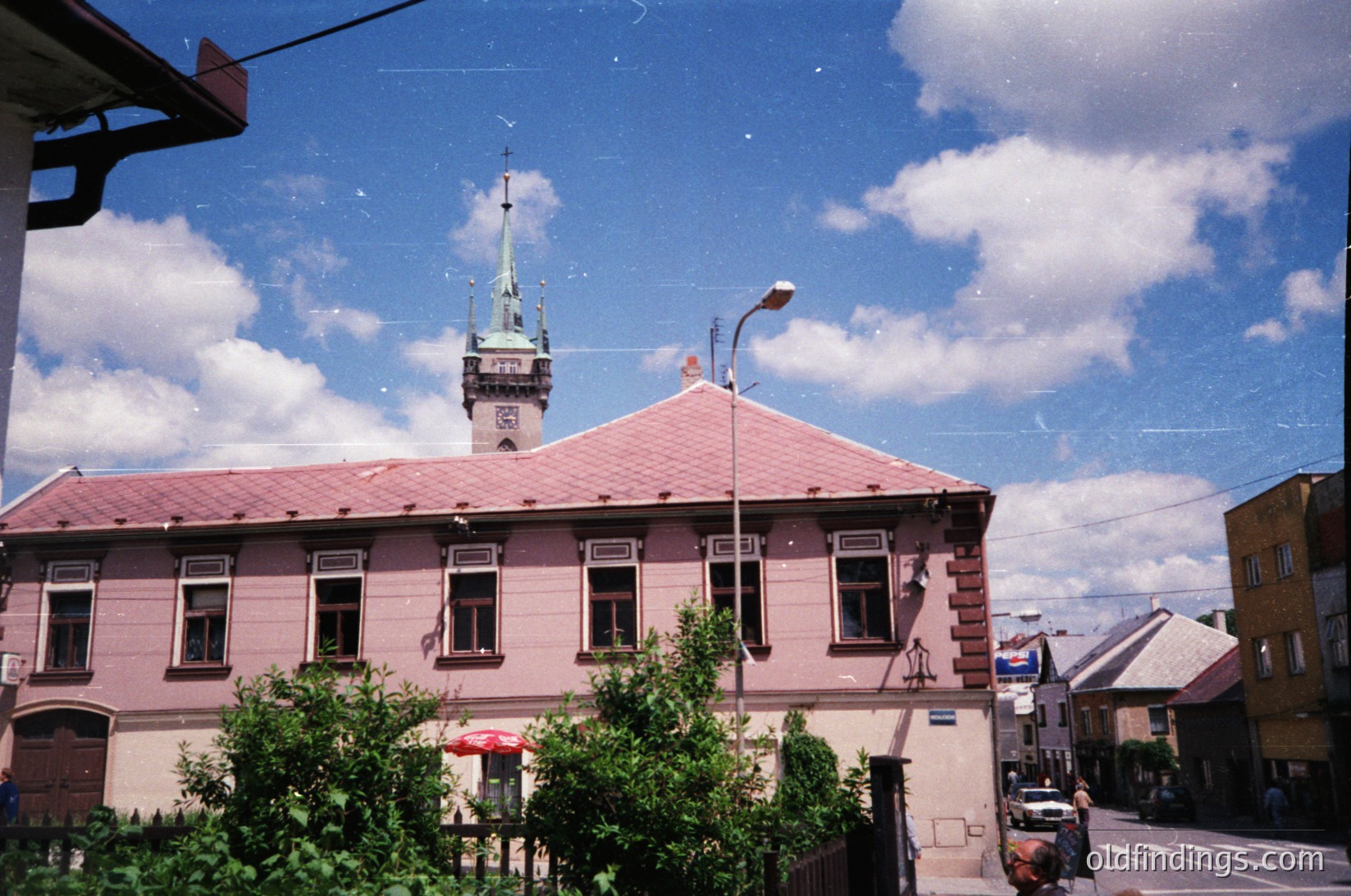 Two-story pink brick building with symmetrical windows, topped by a green-tiled spire and red roof. Urban street scene with vintage street lamp and parked cars. Lush greenery in foreground.