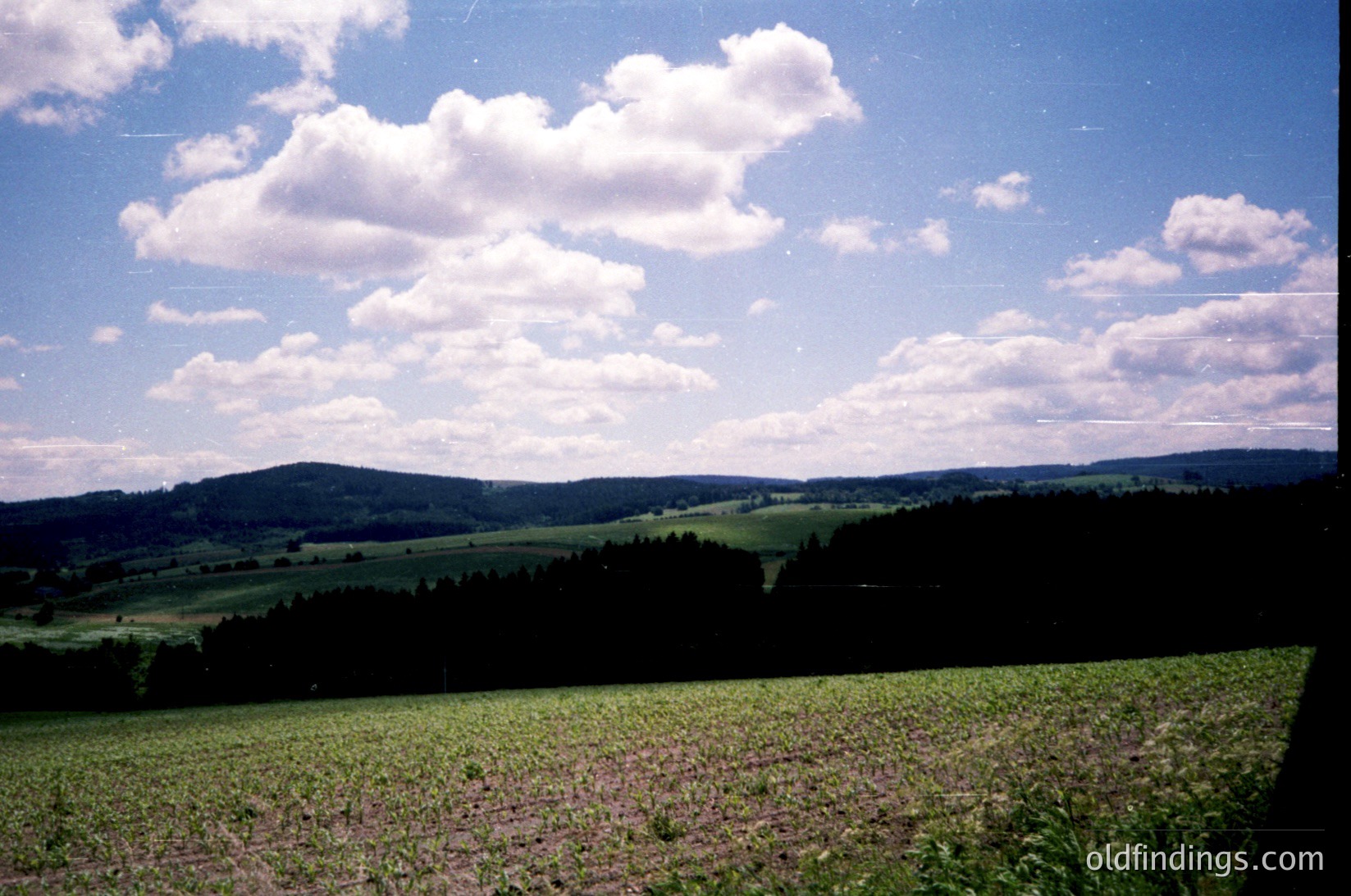 Vintage landscape shot of rolling farmland under bright, fluffy clouds. Green fields stretch toward forested hills under clear skies, suggesting rural agriculture. Color saturation hints at 1970s–1990s film photography.