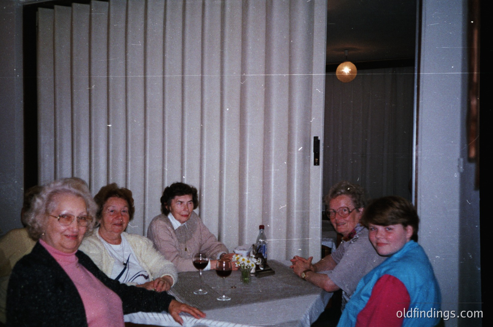 Vintage indoor gathering of five adults seated at a round table in a dimly lit room with vertical blinds. Mid-century retro decor includes a floral-patterned tablecloth and a single pendant light. Glasses, a bottle, and a floral centerpiece suggest a casual meal or celebration. Likely Eastern European setting, 1970s–1980s era.
