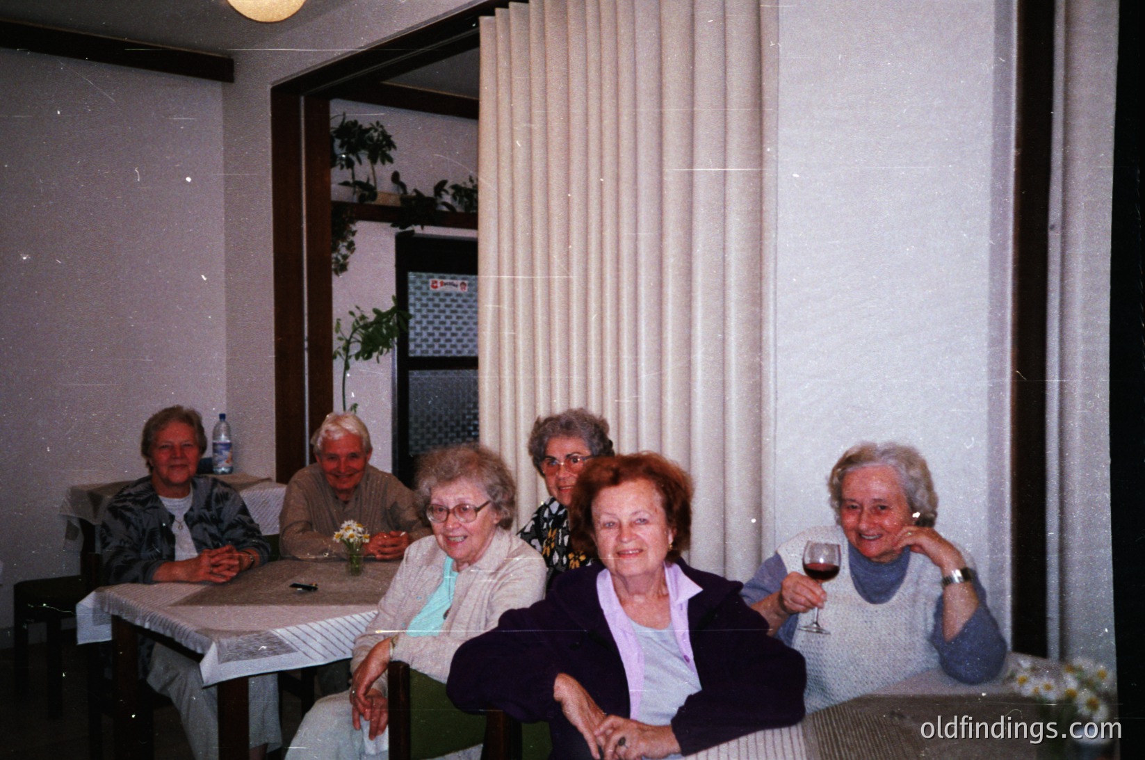 Vintage group portrait of five elderly women in a dimly lit indoor setting, likely a 1980s-1990s European home. Seated at a table with floral tablecloth, they pose with relaxed expressions. One holds a wineglass, another wears a floral necklace. Curtains and a potted plant frame the scene. Authentic mid-century domestic atmosphere.