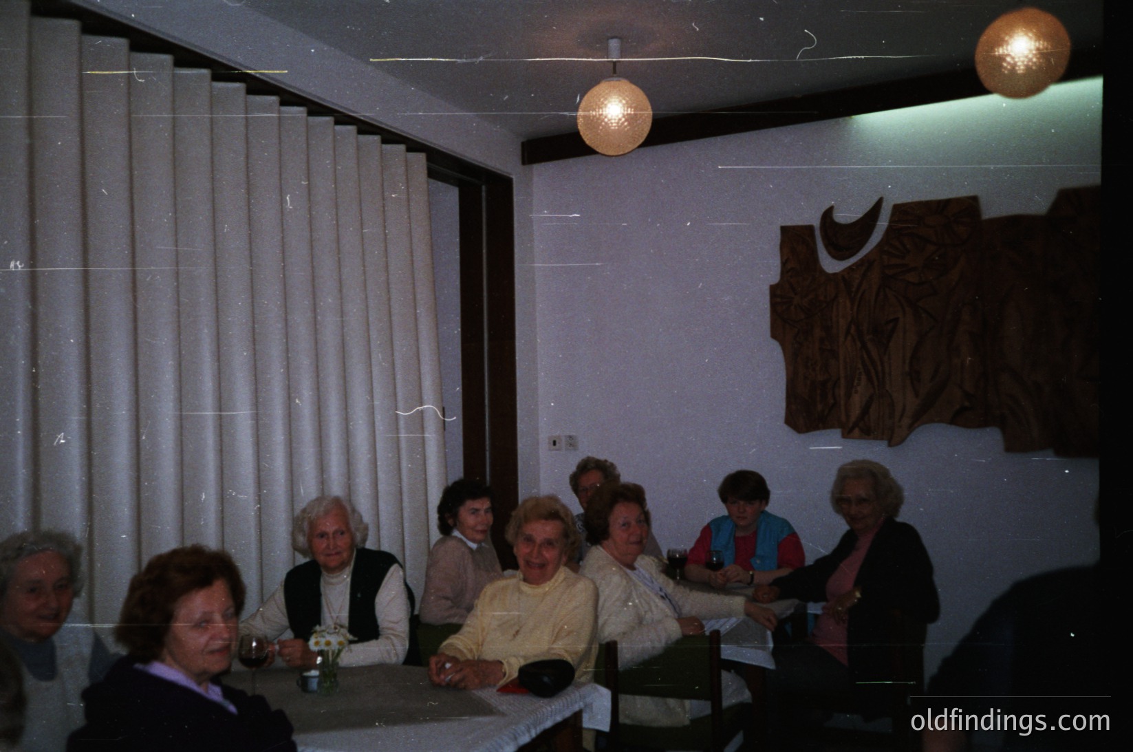 Vintage indoor gathering in a minimalist 1970s-style café or lounge. Eight adults seated around a rectangular table, dressed in layered, patterned clothing. Decor includes hanging spherical lamps, a large abstract wall relief with a crescent moon motif, and vertical blinds. Warm lighting and intimate setting evoke mid-century social culture. éCulture