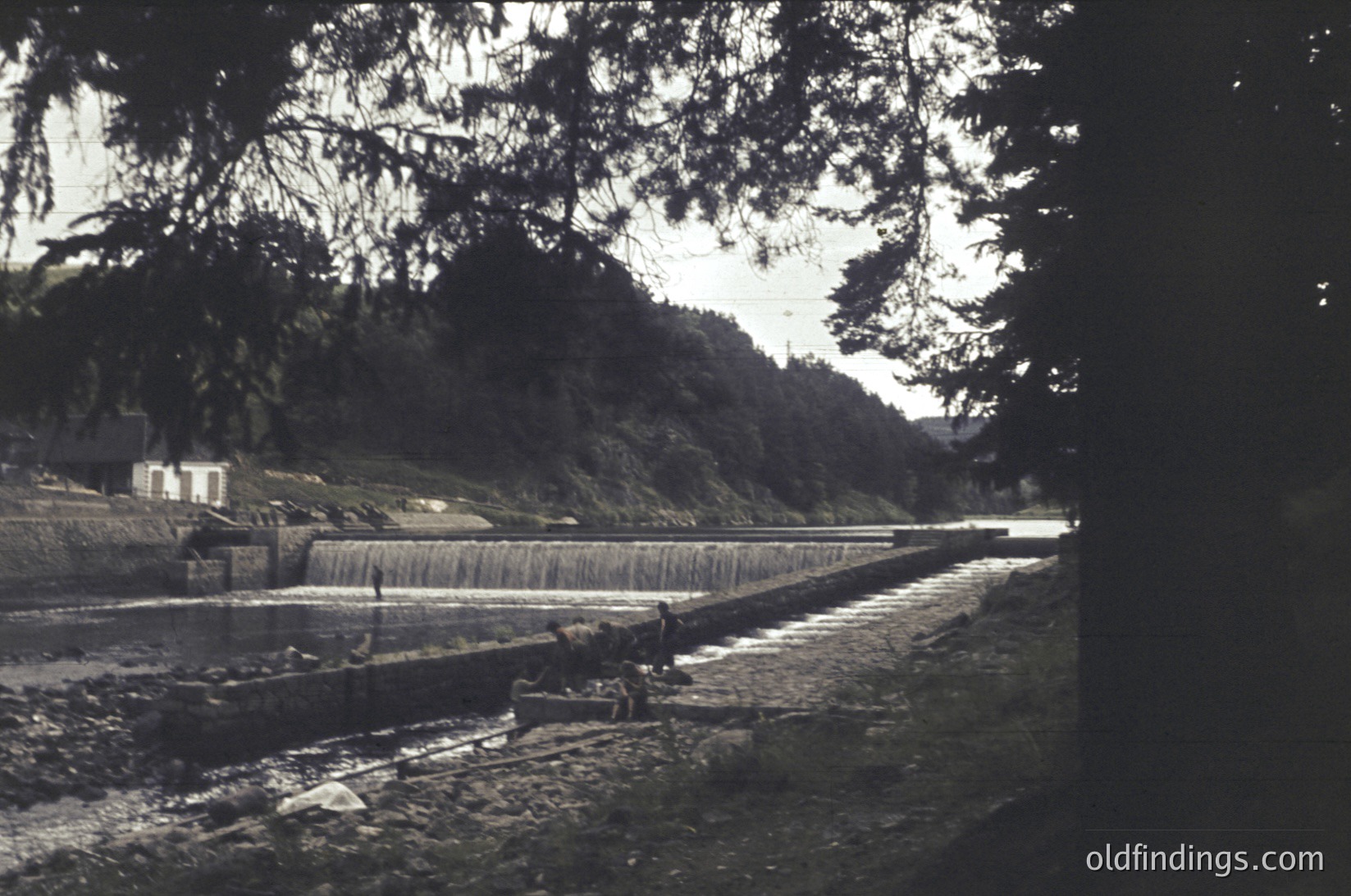 Vintage sepia-toned photo of a small waterfall cascading into a shallow riverbed, framed by dense forest. A lone figure sits on a rock near the water’s edge, possibly fishing or resting. Stone steps and a small structure (likely a cabin or shed) are visible upstream.