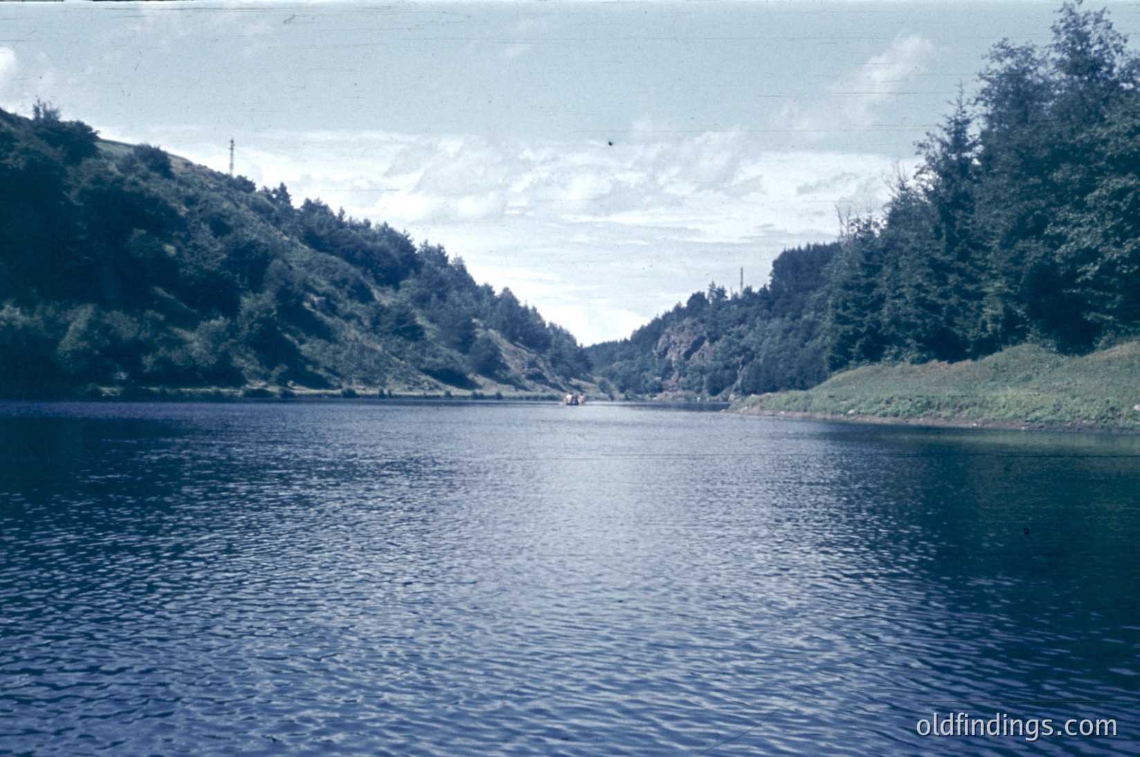 Vintage lake scene with dense forested hills framing calm waters. A lone boat glides near the center, suggesting recreational use. Vibrant greenery contrasts with the muted blue of the water, indicative of mid-20th century color photography.