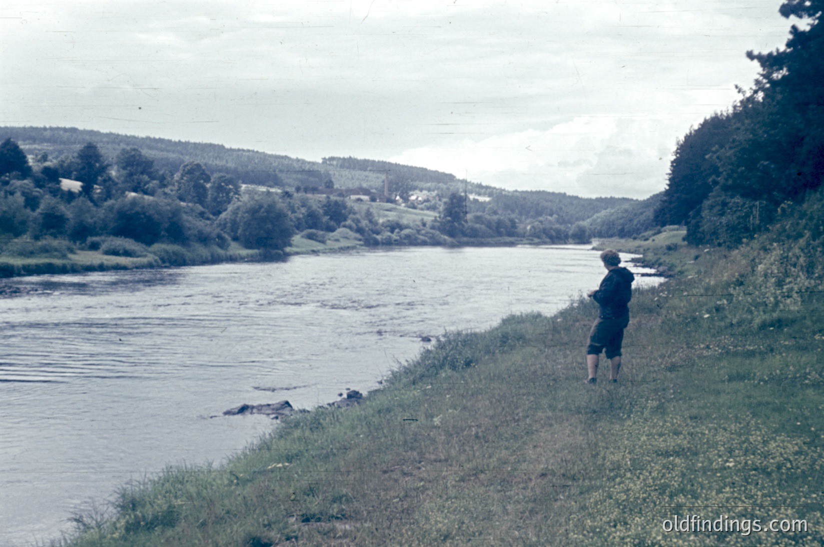 Vintage black-and-white photo of lone fisherman standing on grassy riverbank, holding rod. Wide river curves through lush, forested landscape with distant village. Mid-20th century rural scene.