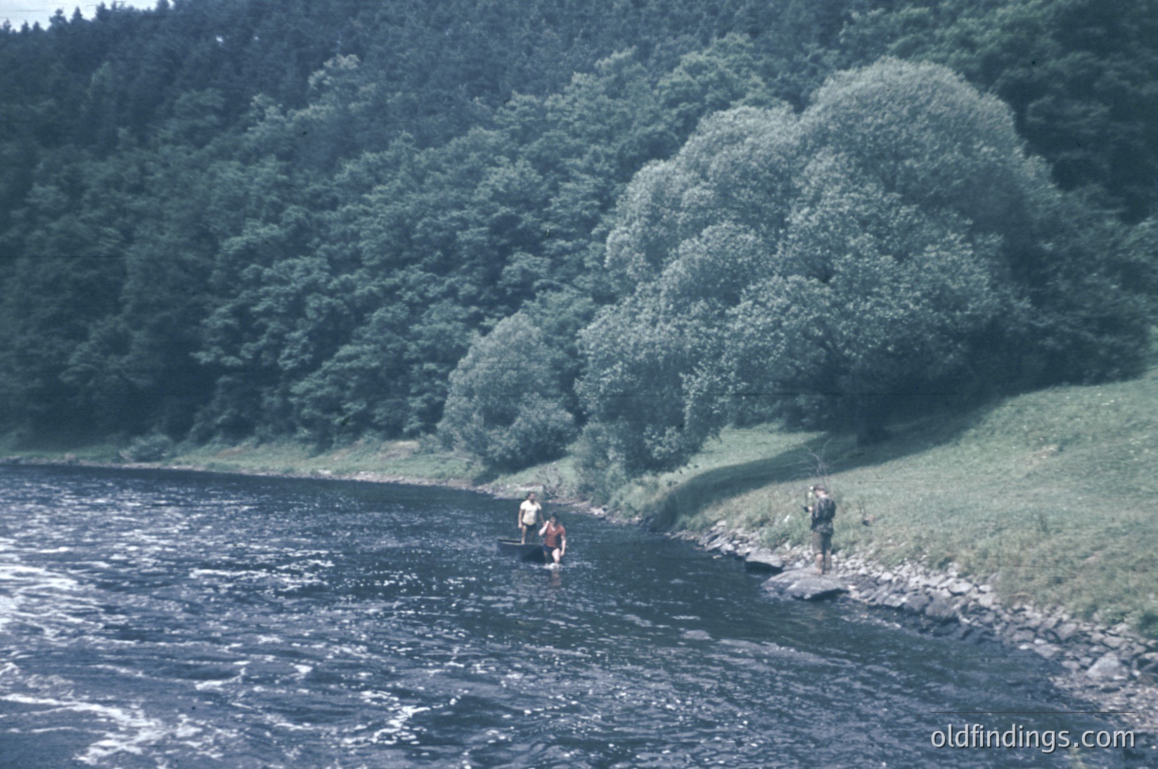 Vintage black-and-white photo of two individuals wading in a shallow, rocky riverbed surrounded by dense forest. One wears a life jacket, holding a fishing rod; the other stands near the water’s edge. Likely mid-20th century outdoor recreation scene.