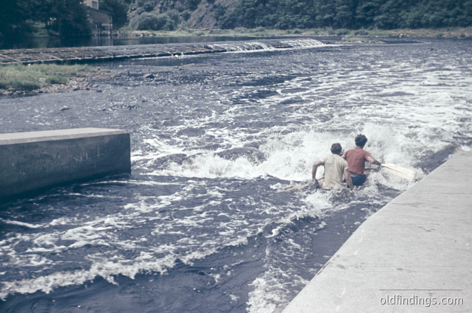 Two young boys wade through turbulent water at a concrete dam spillway, mid-1960s. Vibrant greens contrast with the concrete structure and dark water.