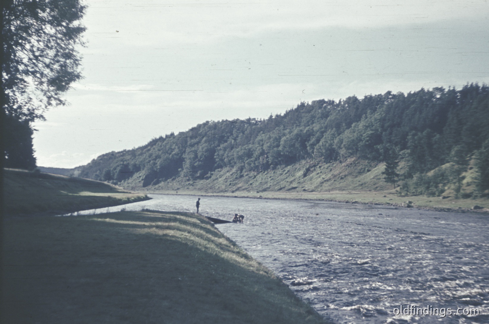 Vintage black-and-white photo of a riverbank scene with lush, forested hills in the background. A lone figure in a boat navigates shallow, turbulent waters near the shore. Mid-20th century aesthetic with grainy texture.