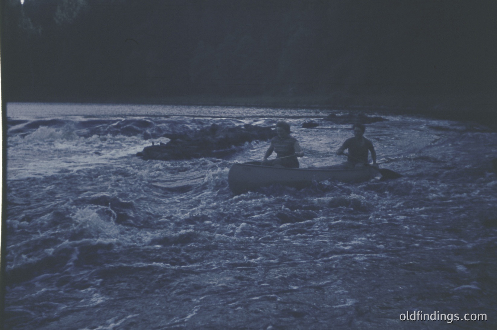 Two individuals paddle a canoe through turbulent waters under low light, likely dawn/dusk. Vibrant blue-green hues dominate the scene, contrasting with the dark silhouette of forested shoreline. Style suggests mid-20th century (1950s–1970s) outdoor recreation.