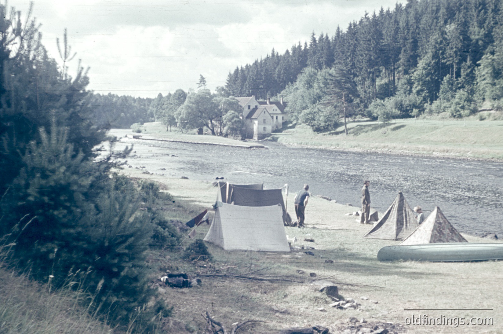 Vintage sepia-toned scene of a riverside campsite with three tents pitched near a small wooden house. Two individuals stand near the tents, one holding a rolled-up sleeping bag. Dense forest and winding road in background. Likely mid-20th century outdoor recreation.