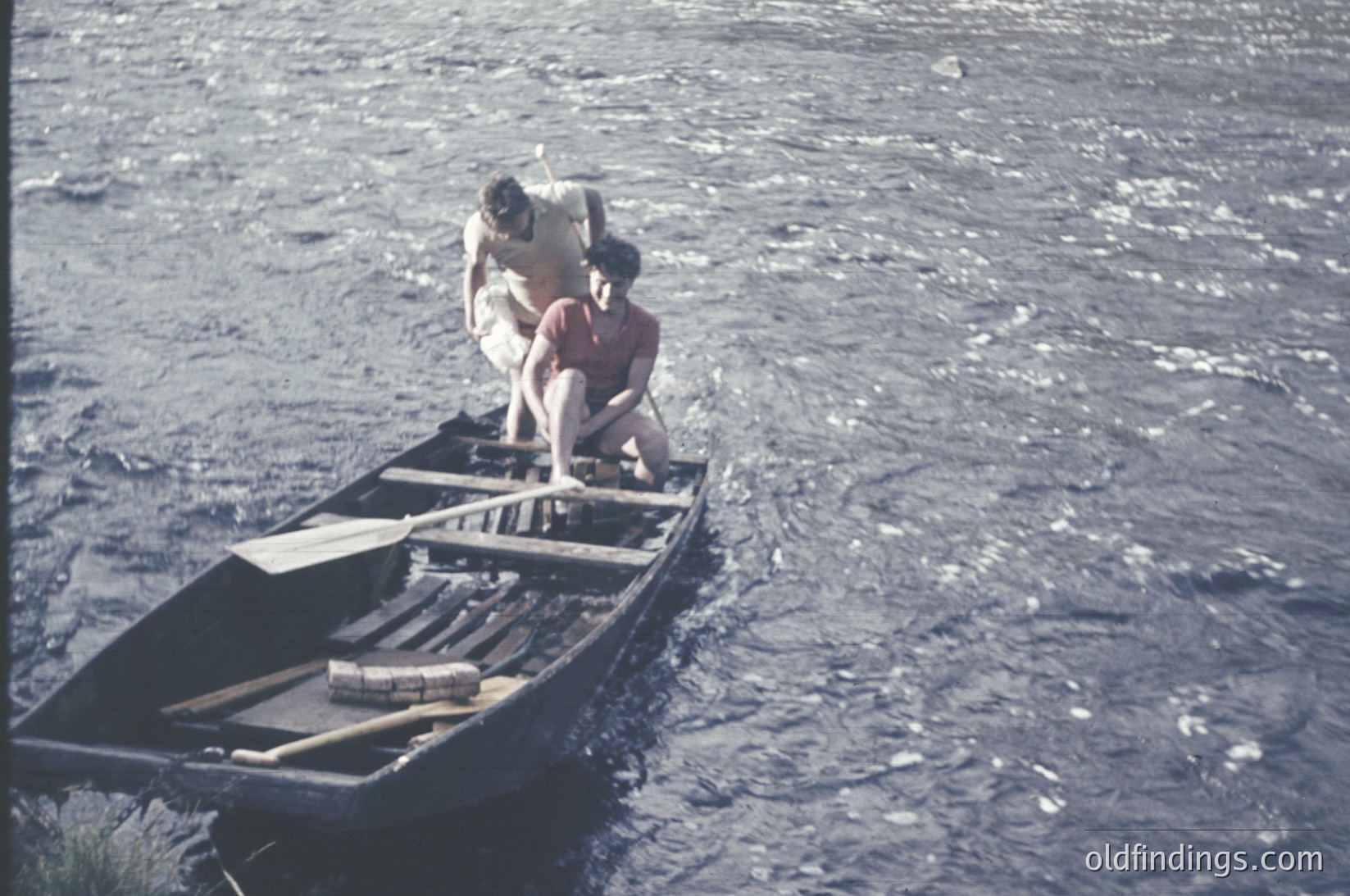 Vintage black-and-white boat scene: two individuals in a narrow wooden canoe, one kneeling to balance while the other stands. Worn wooden paddles and a simple rope tether visible. Likely mid-20th century, possibly . Water appears calm, suggesting a lake or slow river.