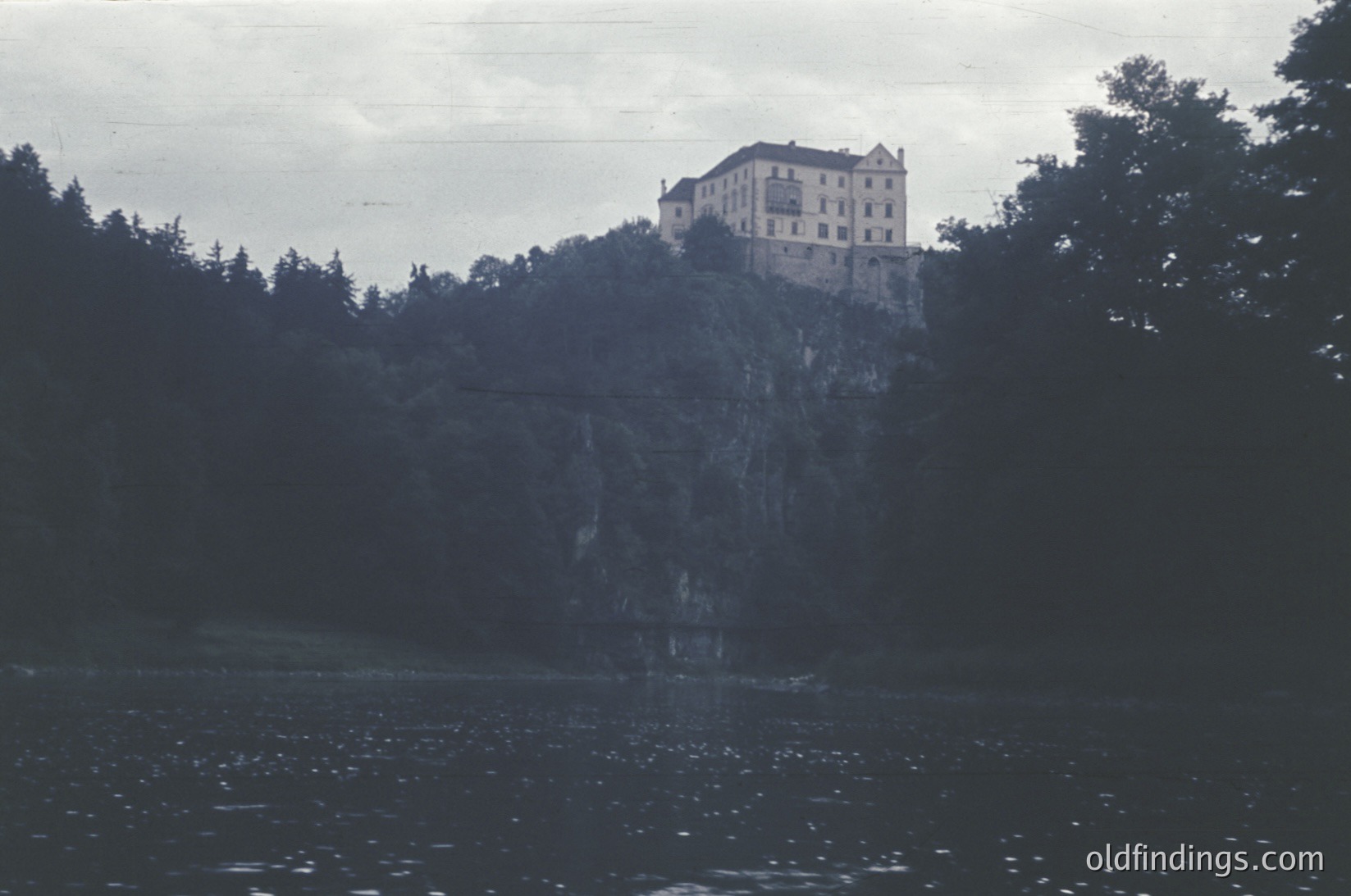 Historic castle perched atop rocky cliffside, surrounded by dense forest. Reflected in calm waters below, suggesting a lakeside or riverbank setting. Architectural style hints at medieval European origins, likely 16th–18th century.