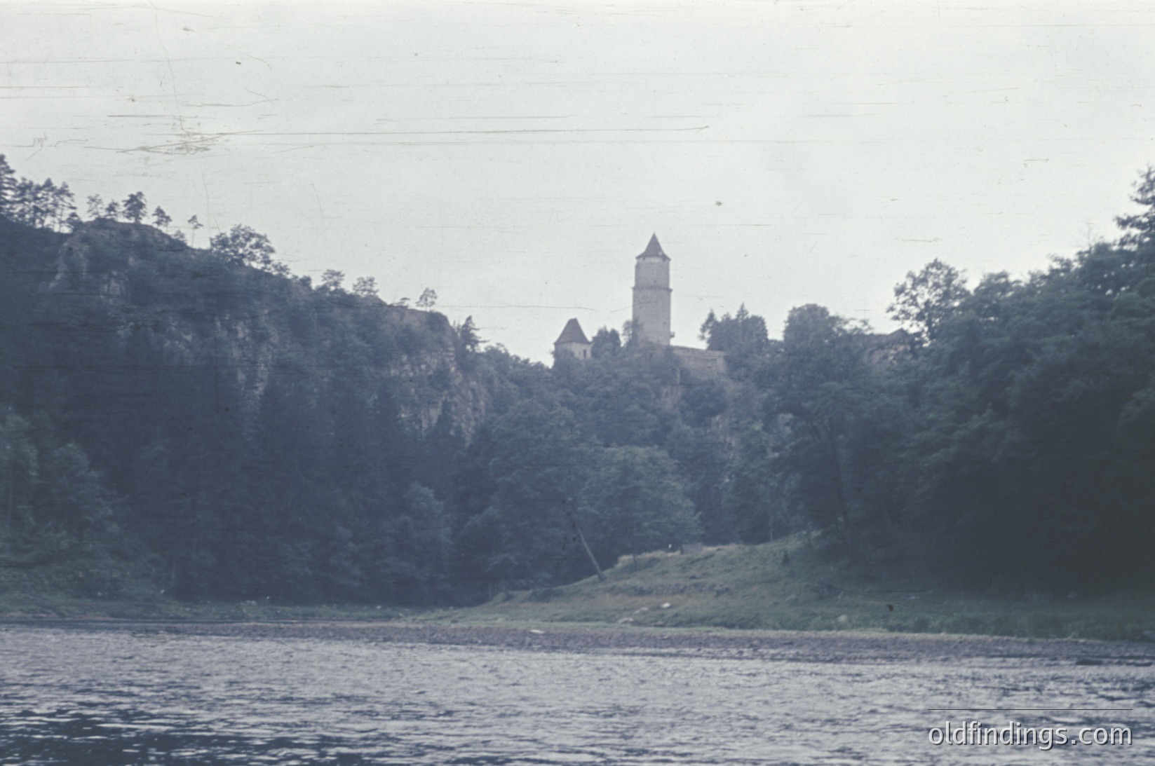 Historic stone castle perched on forested hillside overlooking calm waters, likely a medieval fortress. Architectural details include a prominent round tower and lower defensive walls. Vintage sepia-toned photo suggests early-to-mid 20th century. Potential European location.