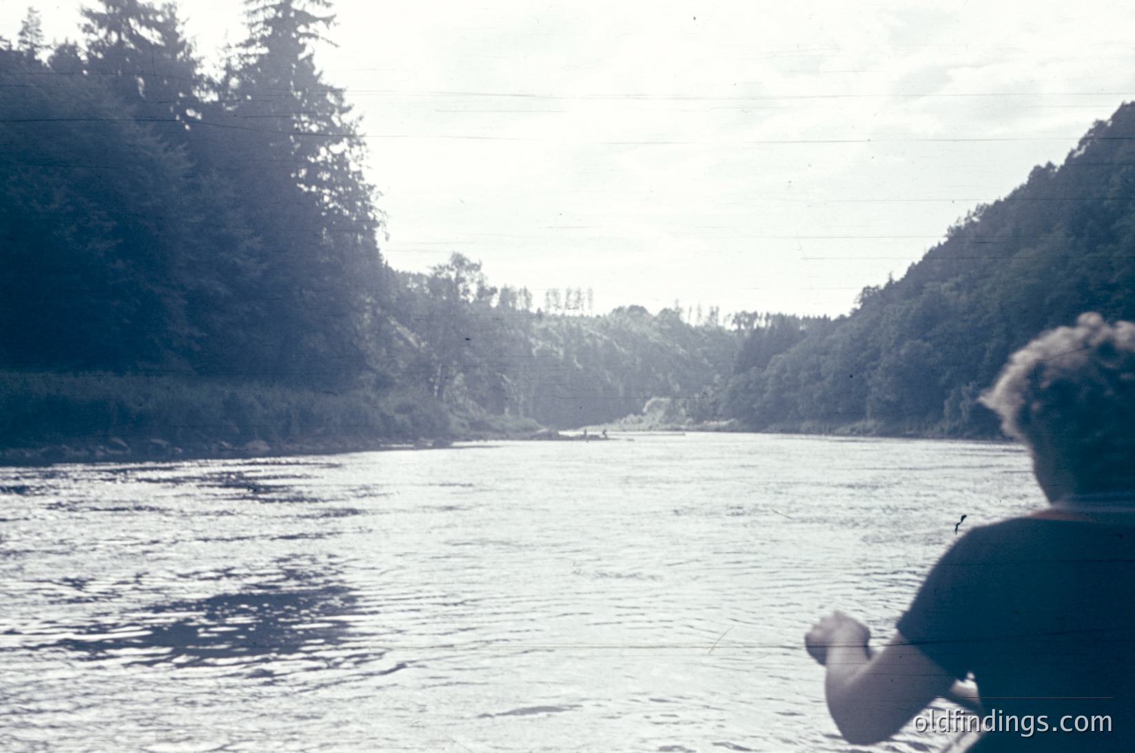 Vintage black-and-white shot of a lone angler casting line into a serene river, flanked by dense forested hills. Overcast sky enhances moody atmosphere. Likely mid-20th century outdoor recreation scene.