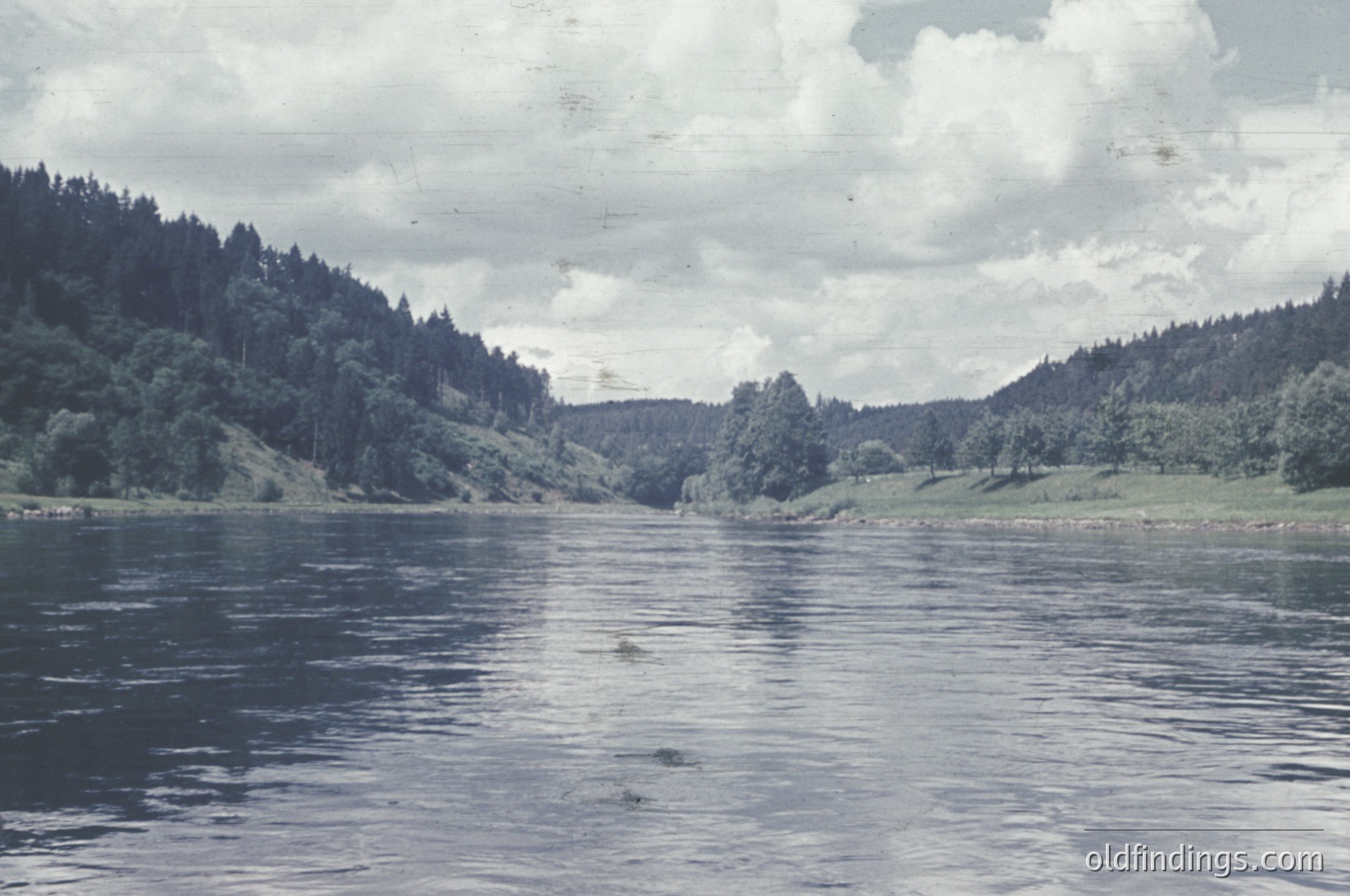 Vintage black-and-white lake scene with dense coniferous forest framing both shores. Gentle ripples reflect muted light on calm waters. Mid-20th century color film tone suggests or . Possible Nordic or alpine region due to forest density and landscape.