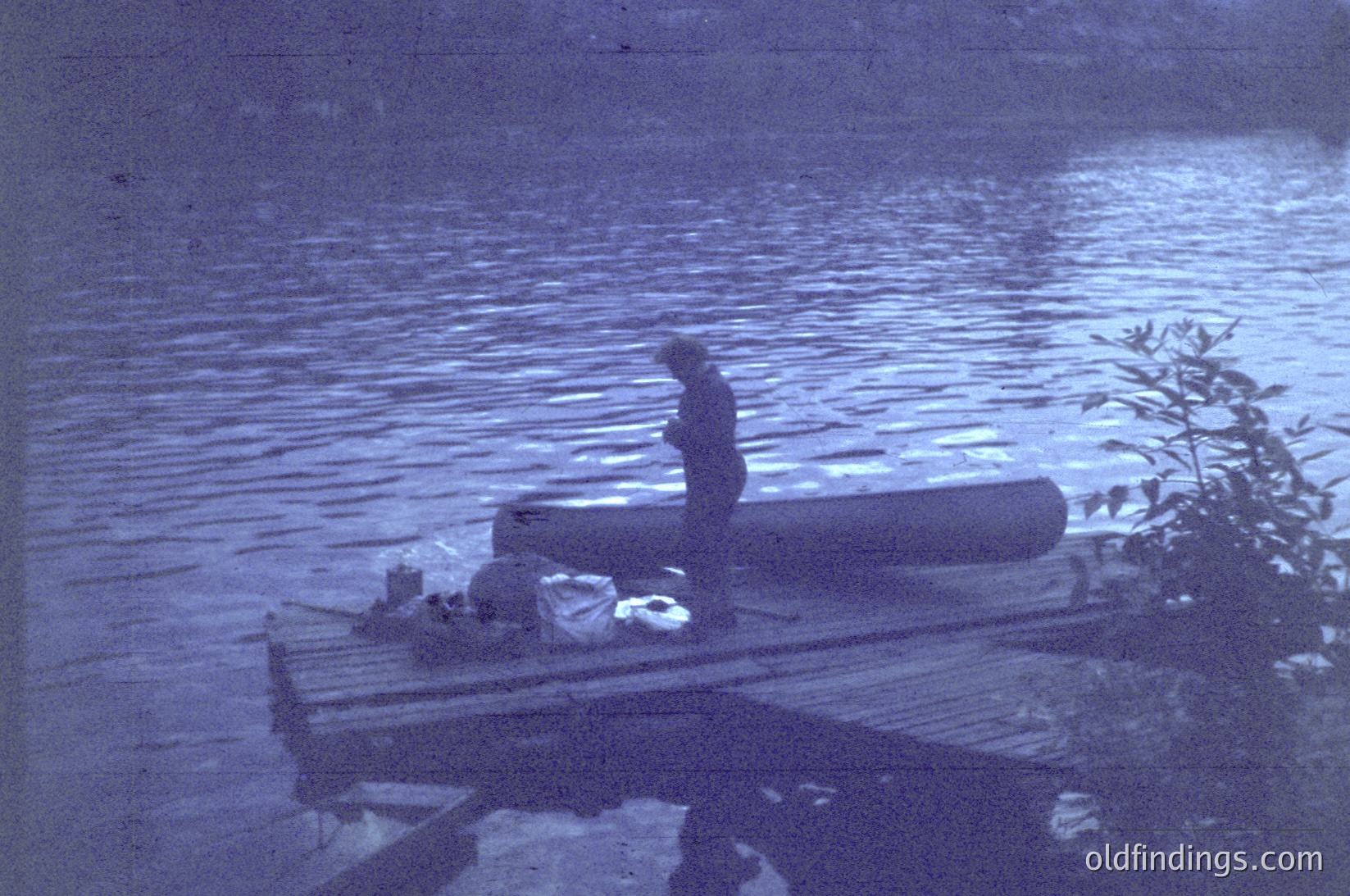 Vintage sepia-toned photograph of a lone figure standing on a weathered wooden dock extending into calm waters. The person appears to be in a contemplative pose, facing the water. The dock is cluttered with miscellaneous items, including a bundle wrapped in fabric. The scene suggests a lakeside or riverside setting, likely from the early-to-mid 20th century.