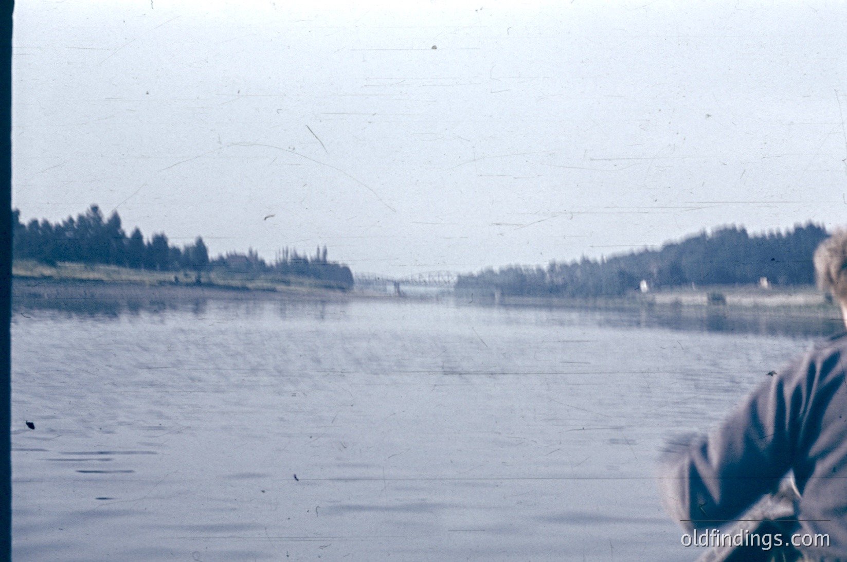 Vintage photo of a serene lake scene with partial view of a person’s arm holding a camera. Dense tree line frames the horizon, suggesting a forested shoreline. Overcast sky and muted tones indicate mid-20th century color film. Likely or outdoor photography.