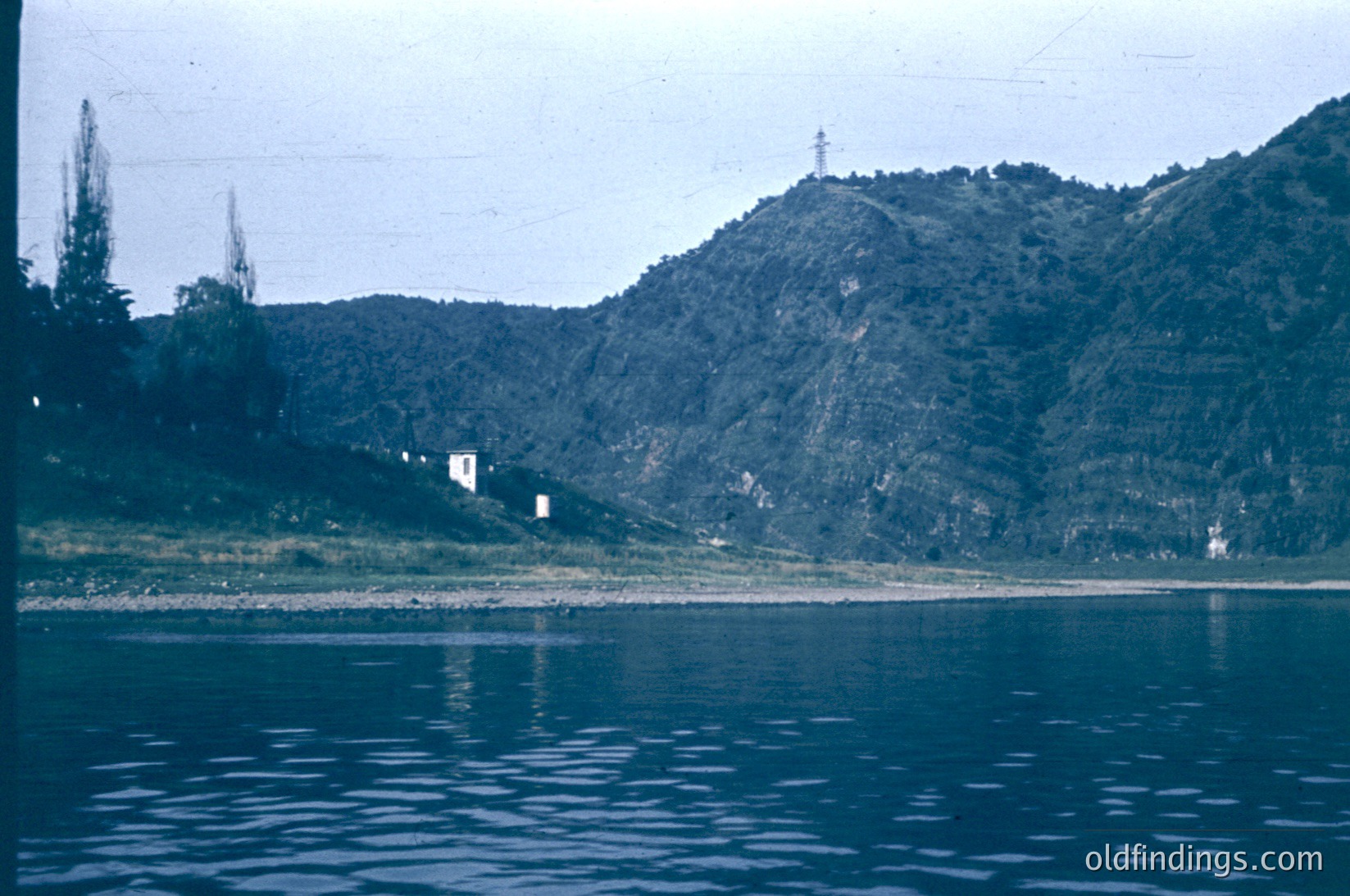 Vintage lake view with forested hills and sparse buildings. The serene reflection in calm waters suggests a remote, natural setting. Likely mid-20th century due to color tone and framing.