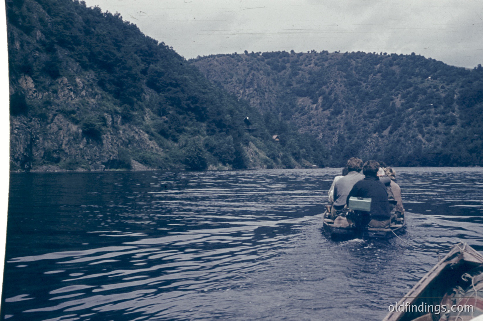 Two scuba divers in full gear float near a rocky coastline, mid-water. Dense forested hills rise steeply in the background. Vintage sepia-toned photo suggests mid-20th century (1950s–1970s) Mediterranean or Adriatic region.
