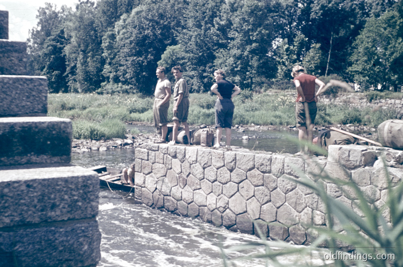 Mid-20th century rural scene: Four individuals in casual summer attire (shorts, blouses) gather near a stone-lined waterway, likely for fishing or leisure. Concrete barriers and cylindrical concrete structures frame the scene. Lush greenery and trees suggest a countryside or lakeside setting.