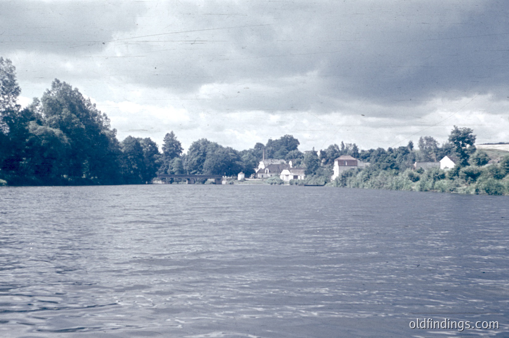 Vintage sepia-toned riverside scene with dense greenery lining both banks. Small, clustered houses with pitched roofs and chimneys suggest a rural European village. Overcast skies and calm water evoke a mid-20th-century atmosphere.
