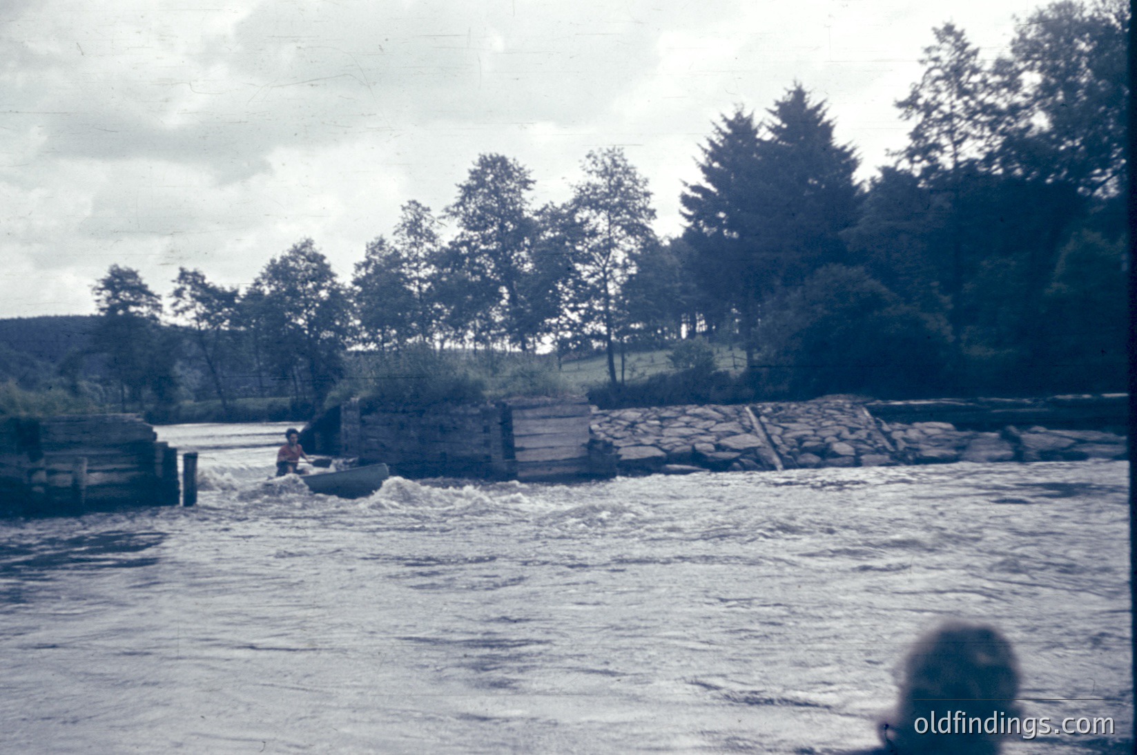 Vintage black-and-white photo of a riverbank with a wooden dock extending into turbulent waters. A lone figure in a boat navigates rapids near a partially submerged concrete structure. Dense forest and overcast skies frame the scene, suggesting a remote, possibly mountainous region. Likely mid-20th century due to grainy film texture.