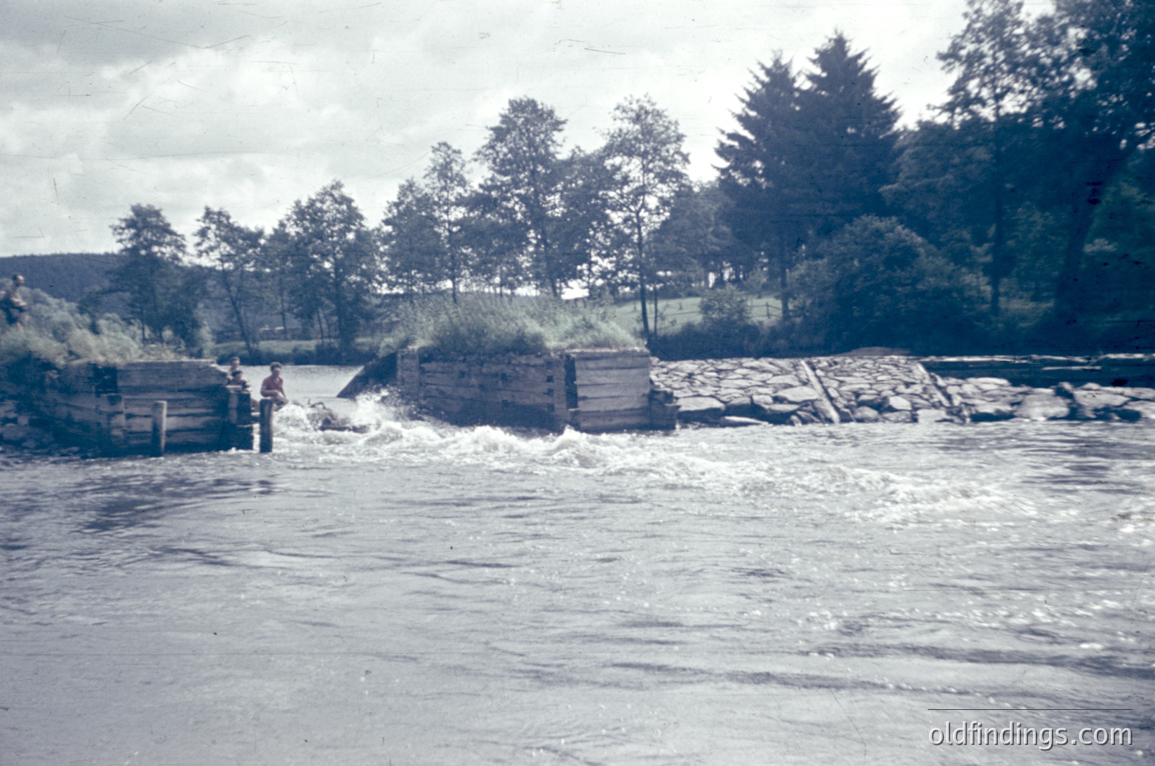 A person wades through a shallow, turbulent river section near a wooden dam or weir, surrounded by lush greenery and trees. The structure appears rustic, likely mid-20th century rural Europe. [Rustic river crossing with wooden dam, lush greenery, mid-20th century ]