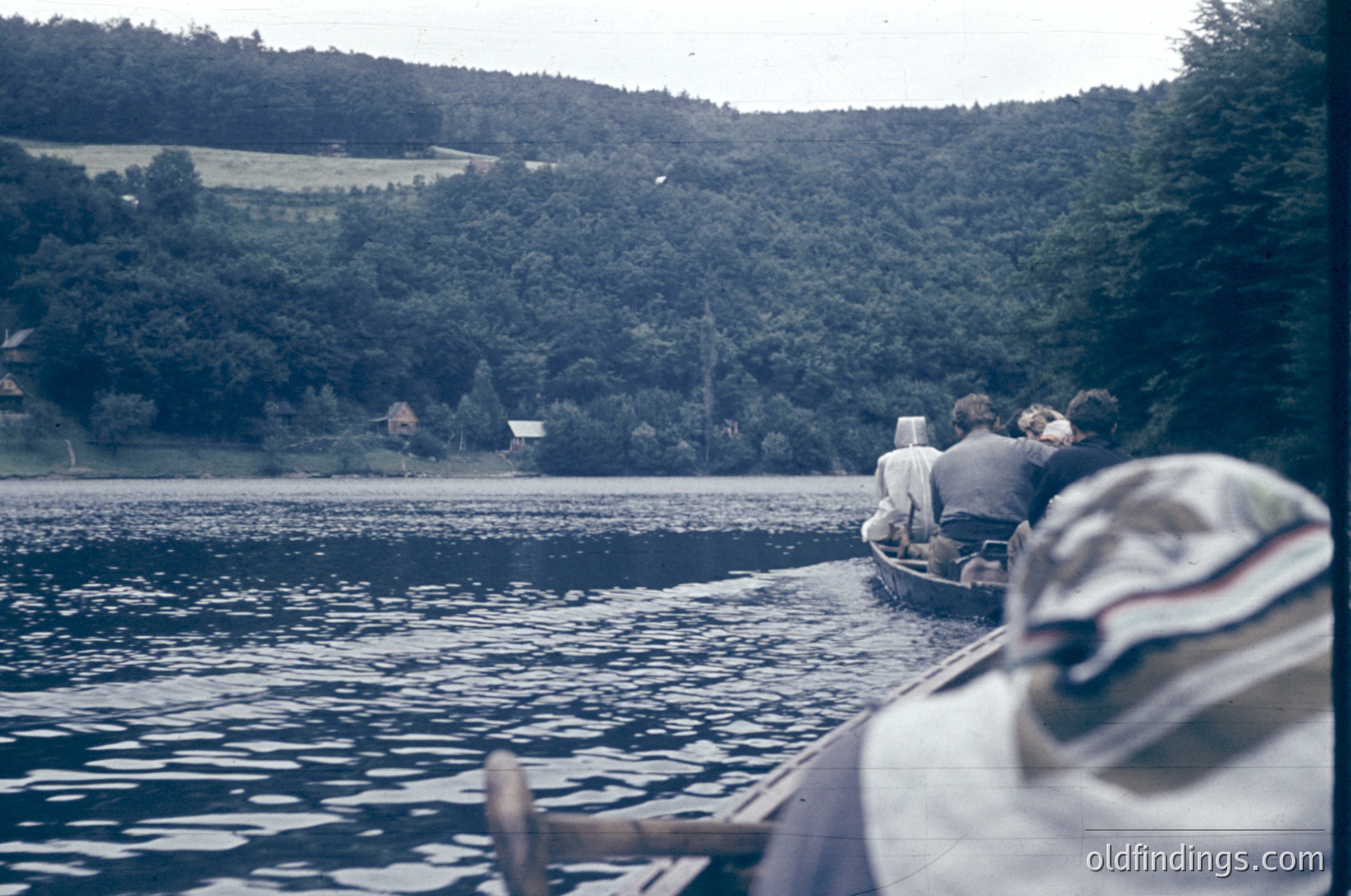 Vintage boat ride on a serene lake surrounded by dense forest and rolling hills. Two individuals in casual summer attire sit in a small wooden boat, paddling toward the shore. The sepia-toned filter suggests a mid-20th century timeframe, likely the 1950s-1960s. Ideal for nostalgic travel or nature-themed content.