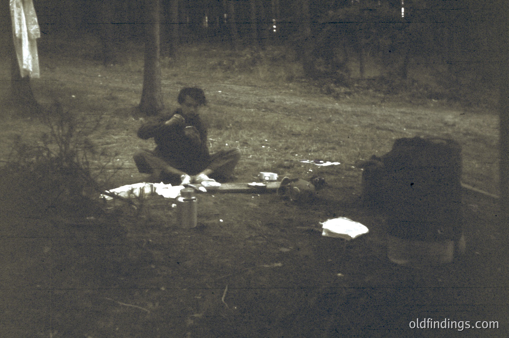 Vintage black-and-white photo of a man seated on grass beside a makeshift campsite at dusk. Notebook and tin can beside him suggest writing or sketching. Surrounding foliage and indistinct background hint at a rural or park setting. Style and lighting suggest mid-20th century.