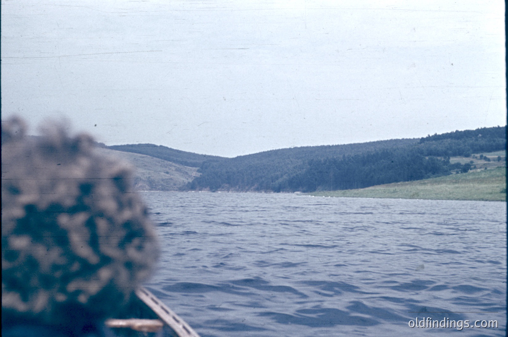 Vintage boat view of a serene coastal landscape with dense forested hills in the background. Water reflects muted blues under overcast skies, suggesting mid-20th century travel photography.