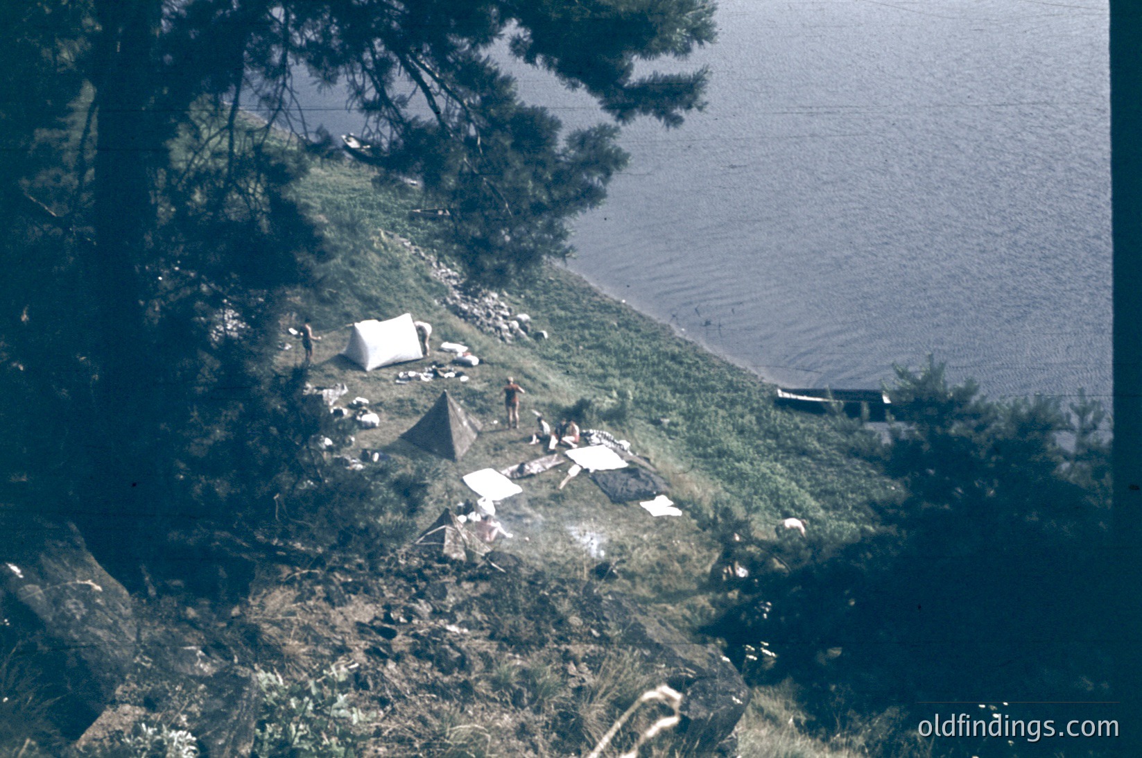 Aerial view of lakeside camping site with tents and scattered gear on grassy, forested shore. Reflective water and a small boat in background. Likely mid-20th century outdoor recreation.