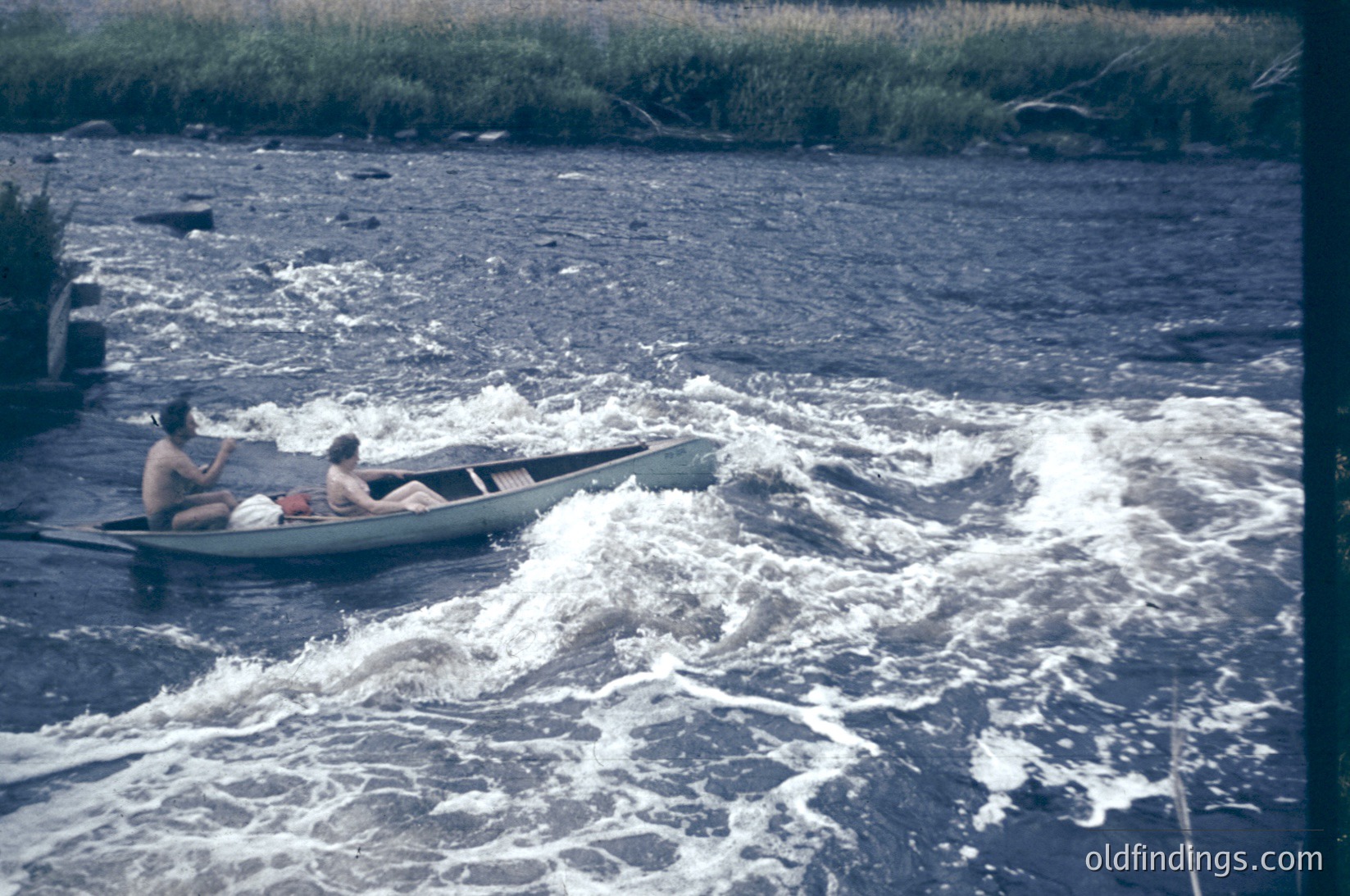 Vintage wooden rowboat navigating turbulent rapids with two seated individuals, likely mid-20th century. Lush reeds and grassy riverbank frame the scene, suggesting a remote or recreational waterway. Color saturation hints at a 1960s–1970s photograph.