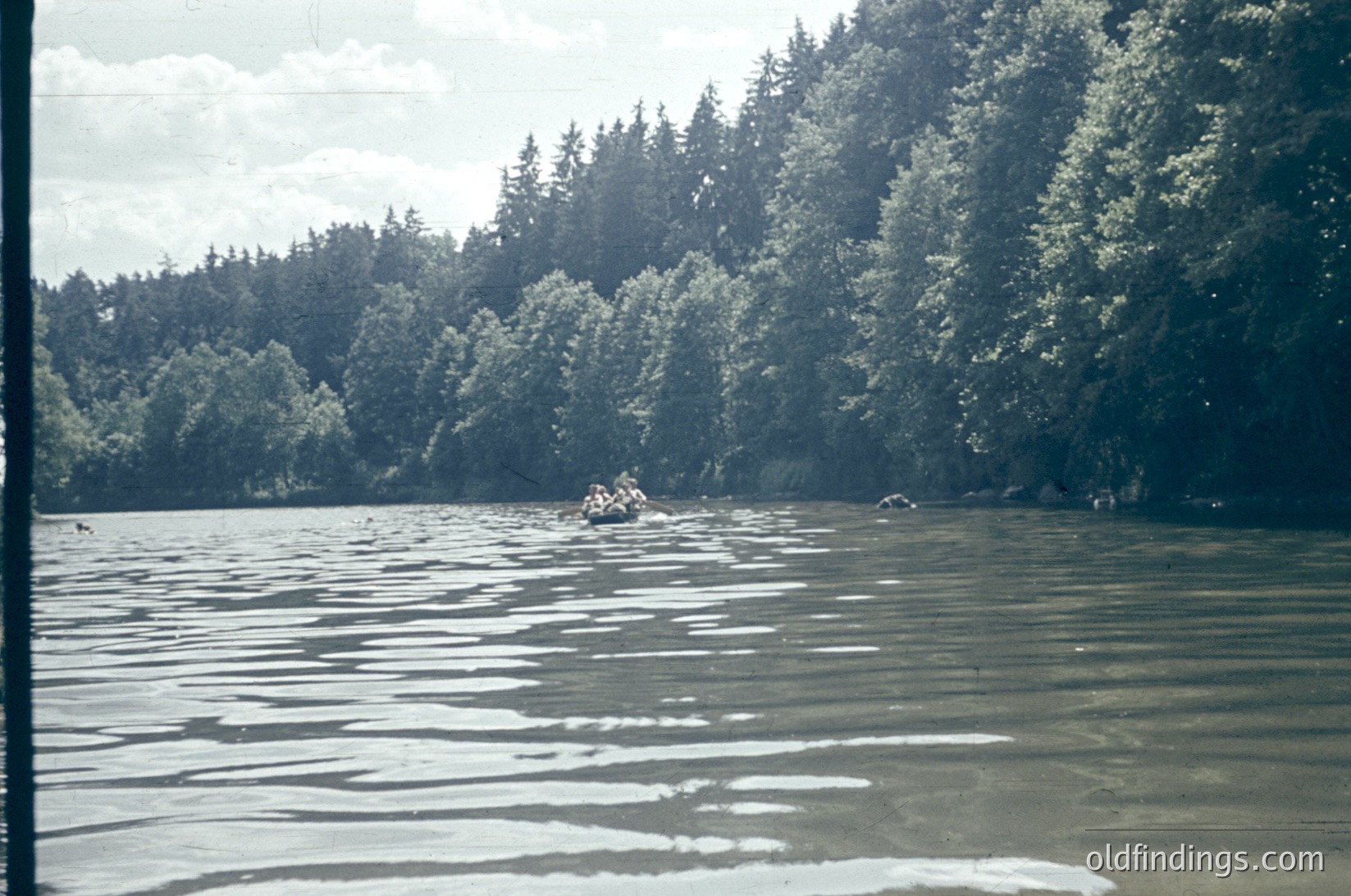 Vintage black-and-white photo of a serene lake surrounded by dense coniferous forest. A small wooden rowboat with two figures paddles near the shore, while another lone figure stands on the water’s edge. The image captures mid-20th century outdoor recreation, likely or . The lush forest and calm water evoke and .