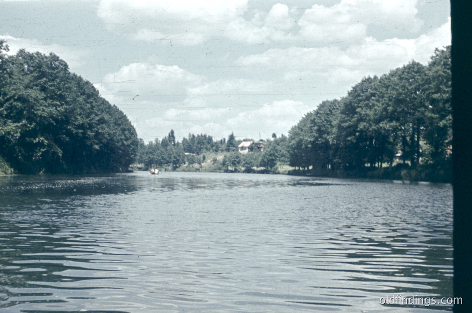 Vintage black-and-white photo of a serene riverbank lined with dense foliage and a single house nestled among trees. The water reflects muted light, suggesting early/late daylight. Likely mid-20th century rural European setting.