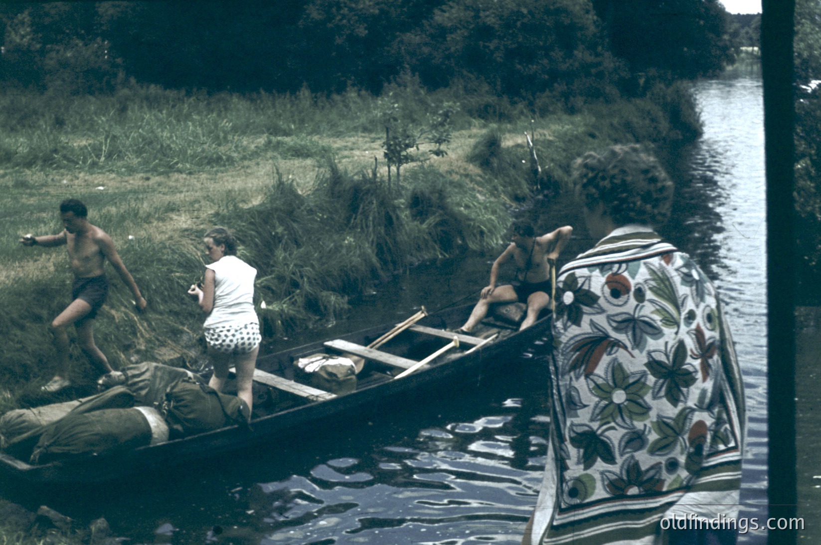 Vintage black-and-white photo (likely 1960s–1970s) of a group launching a wooden canoe into a riverbank. Three people—two men and a woman—position gear and supplies. The woman wears a patterned headscarf and polka-dot swimsuit. Lush greenery and reeds frame the scene.
