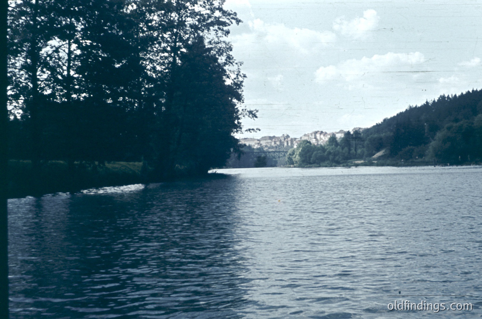 Vintage sepia-toned lake scene with dense forest framing left edge. Distant shoreline shows sparse buildings and forested hills under partly clouded sky. Likely 1950s–1970s European lake region.