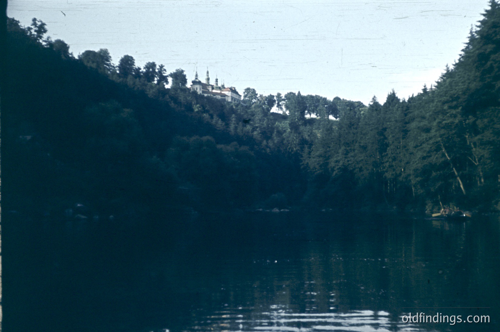 Vintage black-and-white photo of a lakeside scene featuring a grand, multi-domed building perched atop a forested hill. The reflection of trees and structure mirrors on calm water. Likely Eastern European architecture, mid-20th century.
