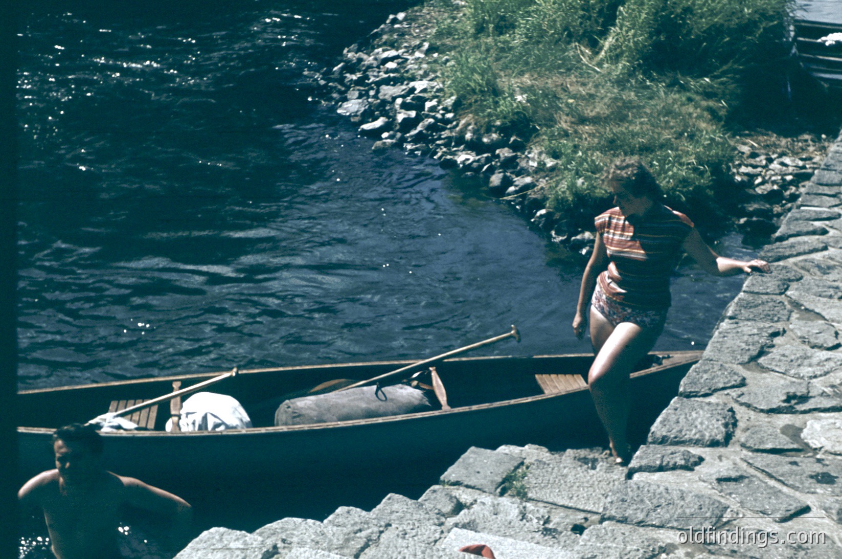 Vintage sepia-toned photo of two individuals preparing a wooden canoe on a stone riverside path. One person stands in the boat, adjusting gear, while another leans over the edge, likely securing equipment. Lush greenery and flowing water frame the scene, suggesting a mid-20th century outdoor adventure.