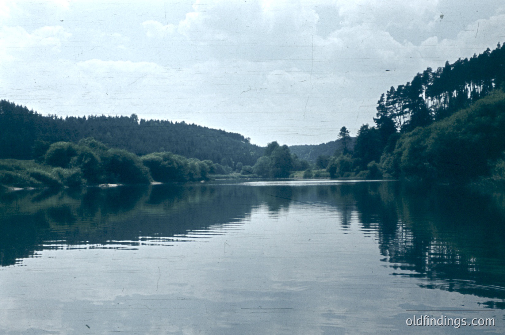 Vintage black-and-white lake scene with dense forest framing both sides, reflecting calm waters. Overcast skies enhance serene, muted tones. Likely mid-20th century due to film grain and style.