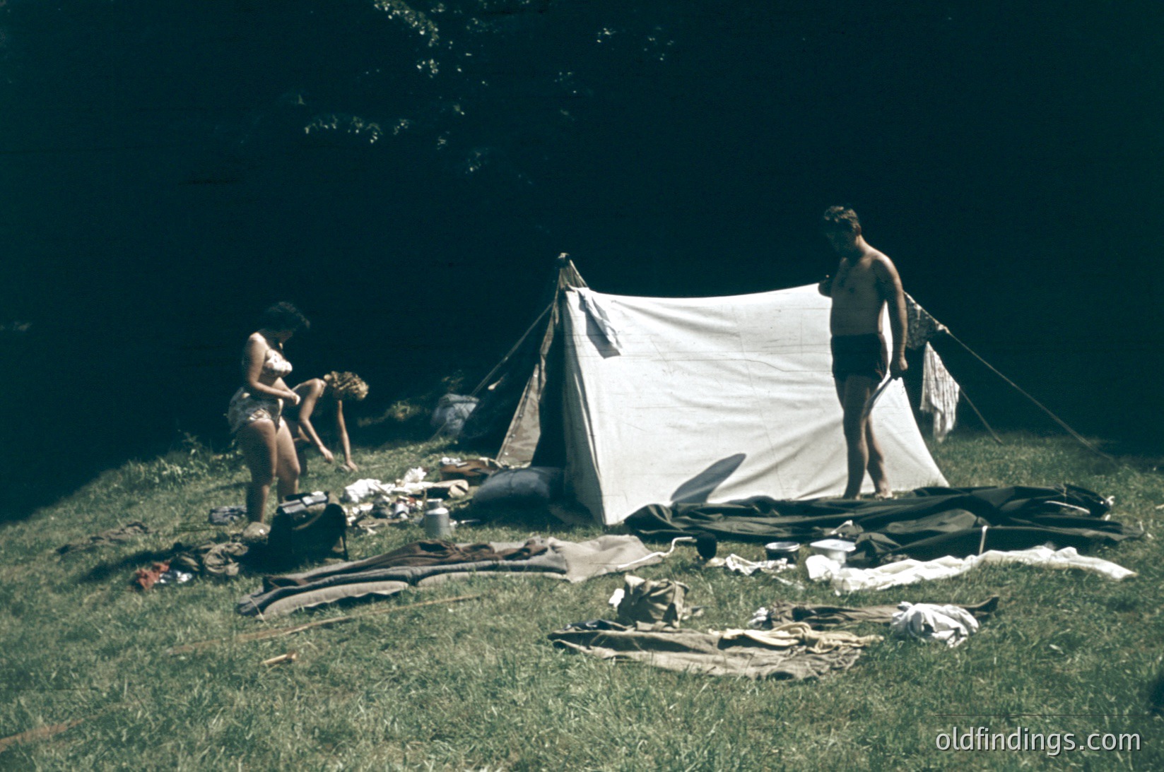 Vintage camping scene with two individuals setting up a white canvas tent in a grassy, wooded area. One person kneels organizing gear, while the other stands shirtless, holding tent poles. Surrounding equipment includes rolled sleeping bags, a blanket, and a backpack. Warm, sepia-toned photo suggests 1960s–1970s outdoor recreation.