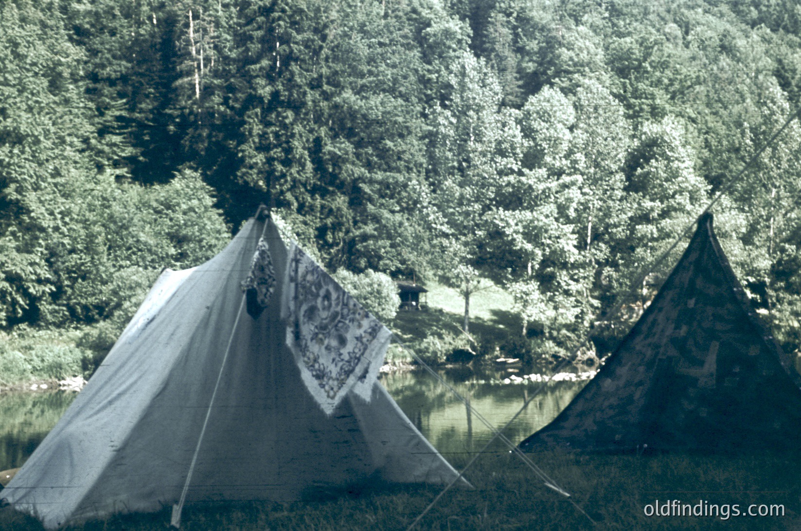 Vintage-style tents pitched beside a serene lake, surrounded by dense coniferous forest. The tent on the left features a floral-patterned mesh panel, while the right tent displays a geometric design. Reflections in the water enhance the tranquil alpine setting. Likely mid-20th century outdoor recreation.