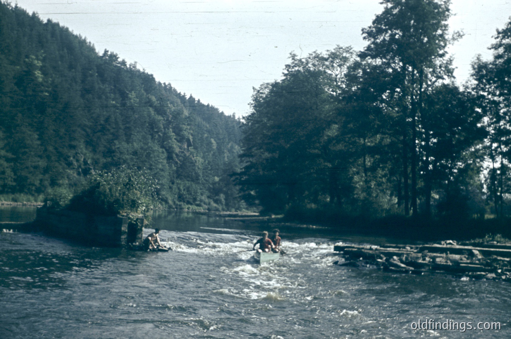 Vintage photo of three kayakers navigating a rapid in a forested river, likely mid-20th century. Lush green hills and dense pine trees frame the scene, with a wooden bridge or dock visible on the right. Water appears turbulent, suggesting outdoor adventure activity.