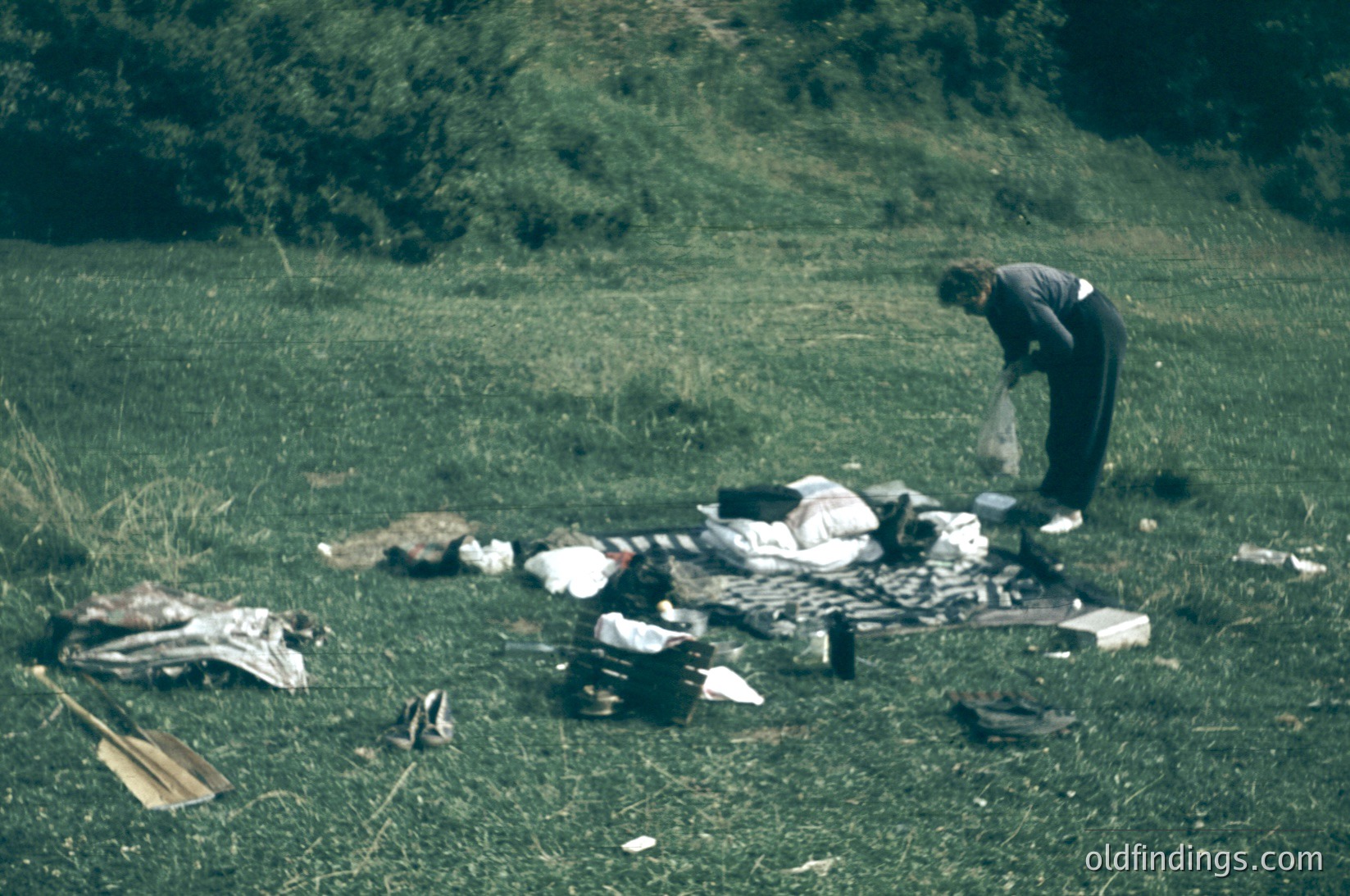 Mid-century outdoor picnic scene with vintage clothing and camping gear. A person in a striped sweater and dark pants organizes a spread of folded blankets, a striped towel, and a wooden paddle. Lush green hillside and grassy field suggest a rural or park setting. Likely 1950s–1970s.