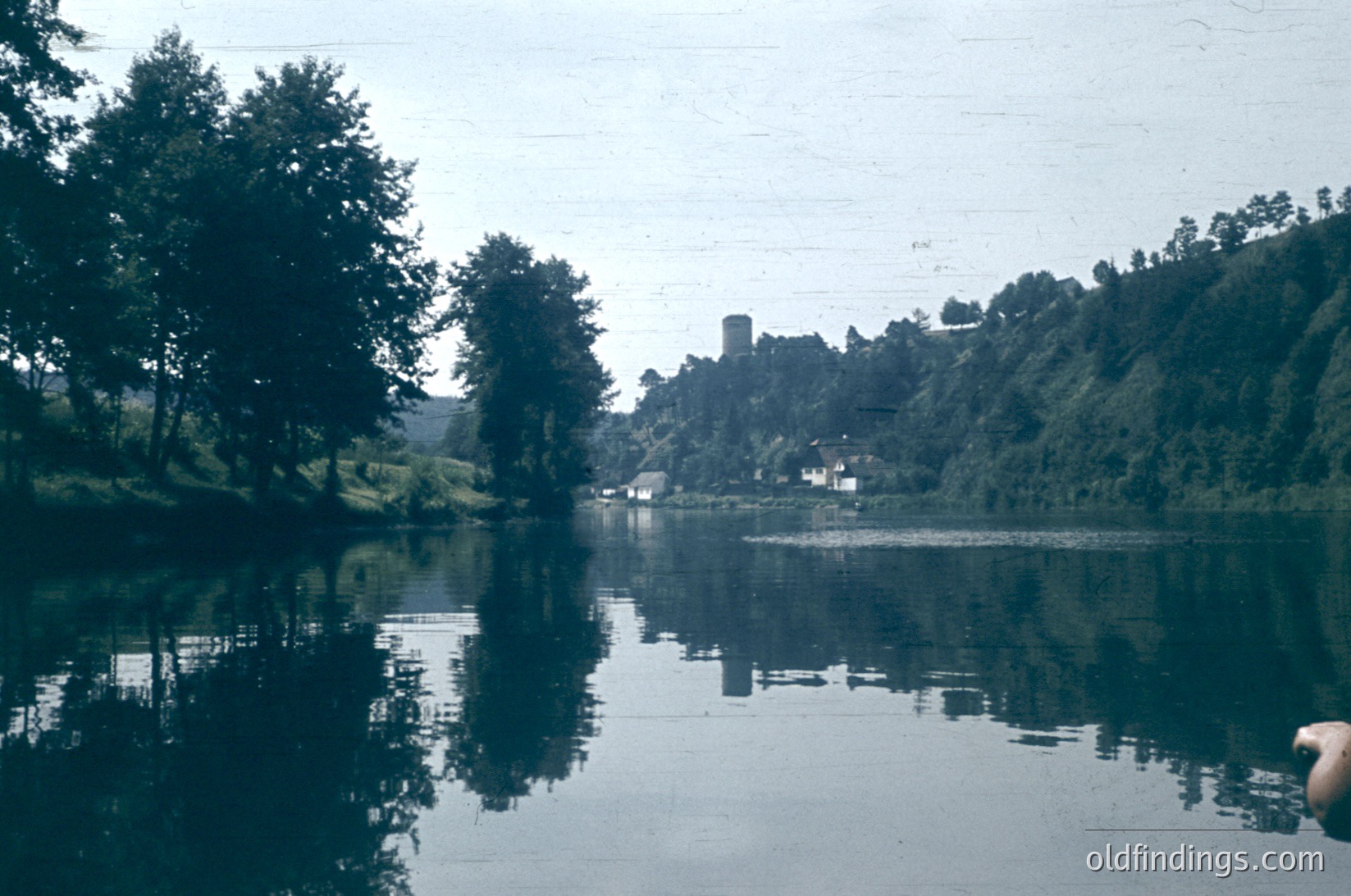 Vintage black-and-white lake scene with a medieval stone tower partially obscured by dense foliage. Reflections create mirror-like calmness. Rural European village with thatched roofs and scattered buildings along shoreline. Likely 19th-century European countryside.