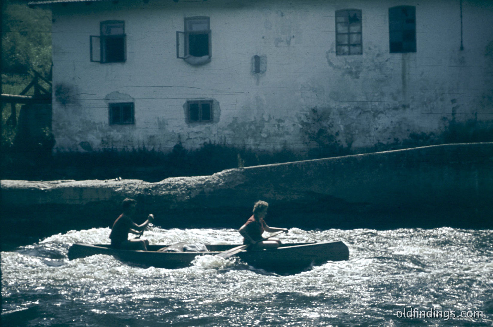 Two individuals in a narrow wooden boat navigate turbulent river waters, likely for fishing or transport. Behind them, a weathered concrete building with rectangular windows and exposed wiring suggests rural or industrial use. The scene evokes mid-20th century rural life.