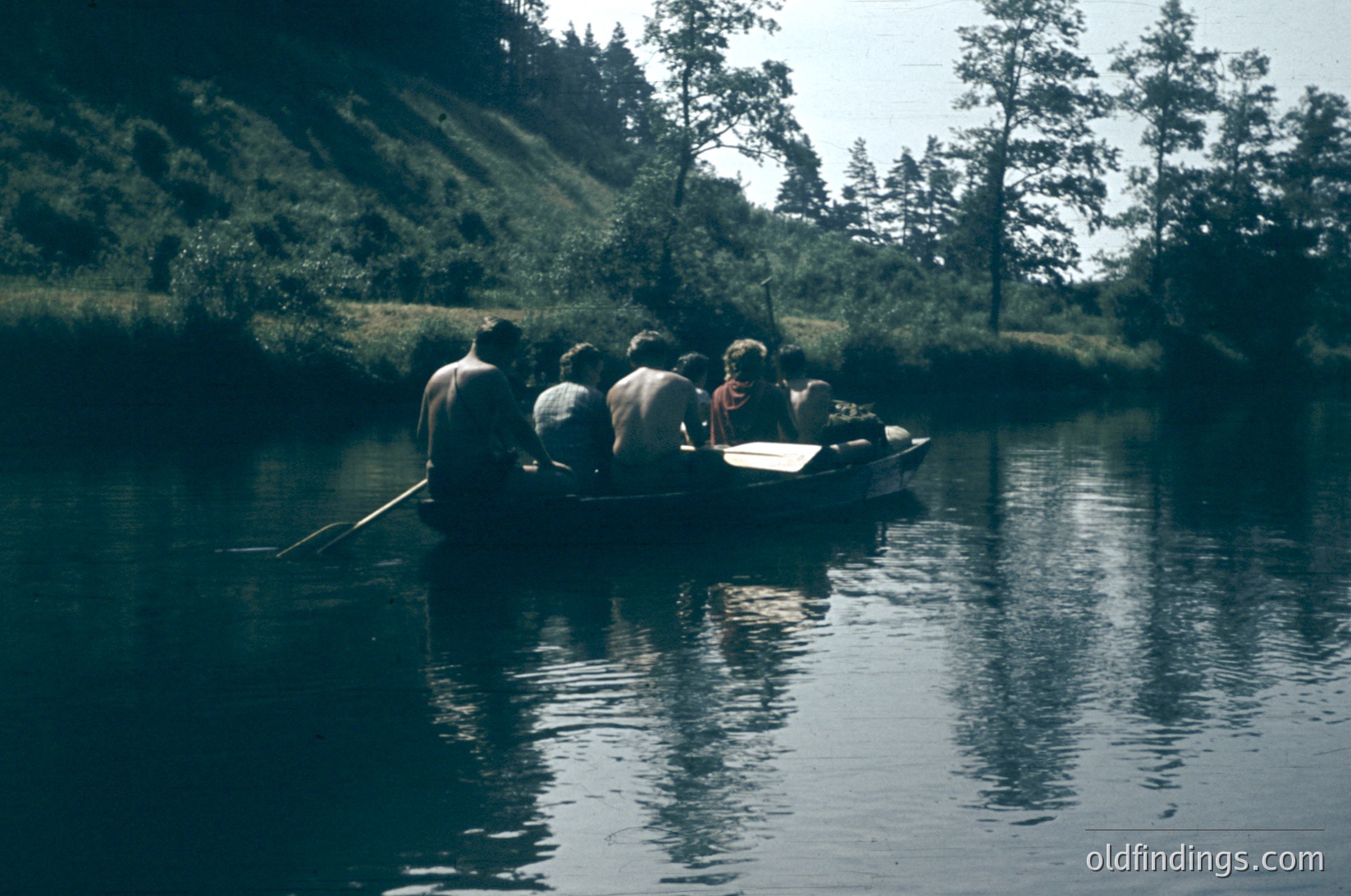 Six individuals in a narrow wooden boat on a calm river, paddled by one person at the rear. Lush, forested hillsides frame the scene, suggesting a rural or wilderness setting. Sepia-toned, likely from the mid-20th century.
