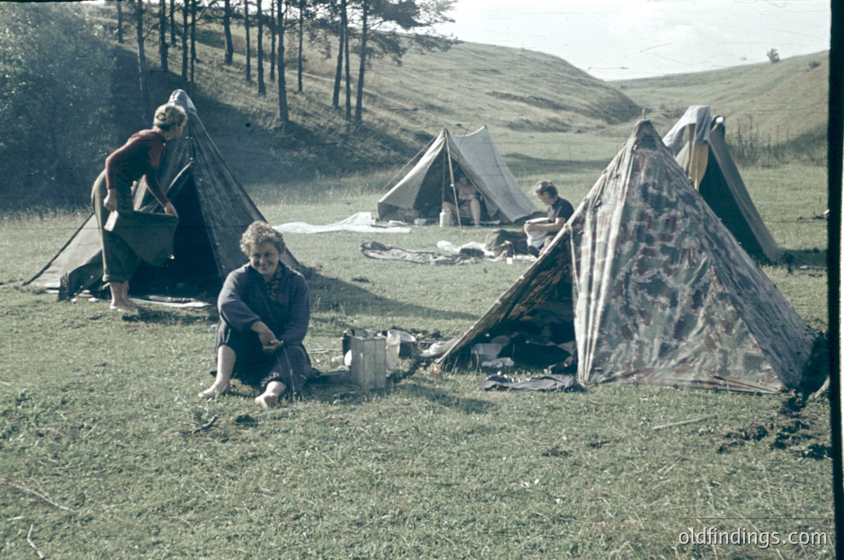 Vintage camping scene in a grassy meadow with three canvas tents. Two women—one standing near a cooking pot, the other seated with a child—suggest mid-20th-century outdoor recreation. Surrounding hills and forest indicate a rural, possibly European setting.