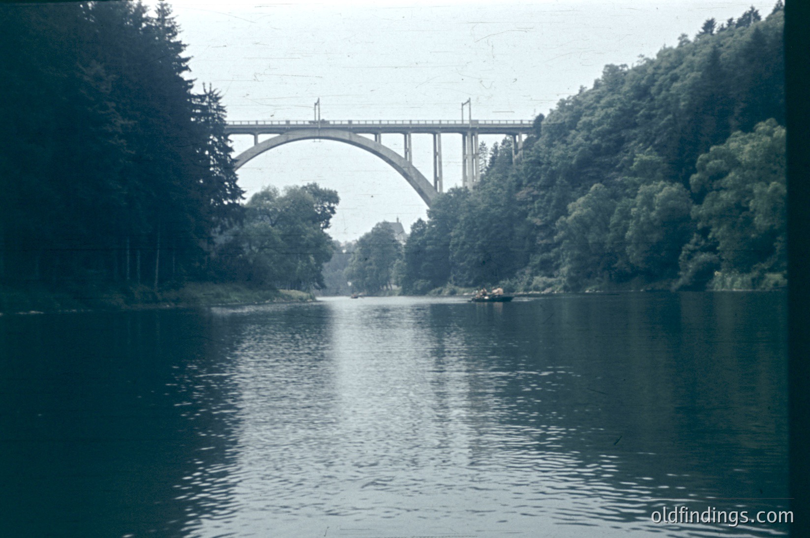 Mid-20th century concrete arch bridge spanning a calm river, flanked by dense forest. Symmetrical design with two prominent arches. Vintage sepia tone suggests or era. Potential industrial or rural setting.