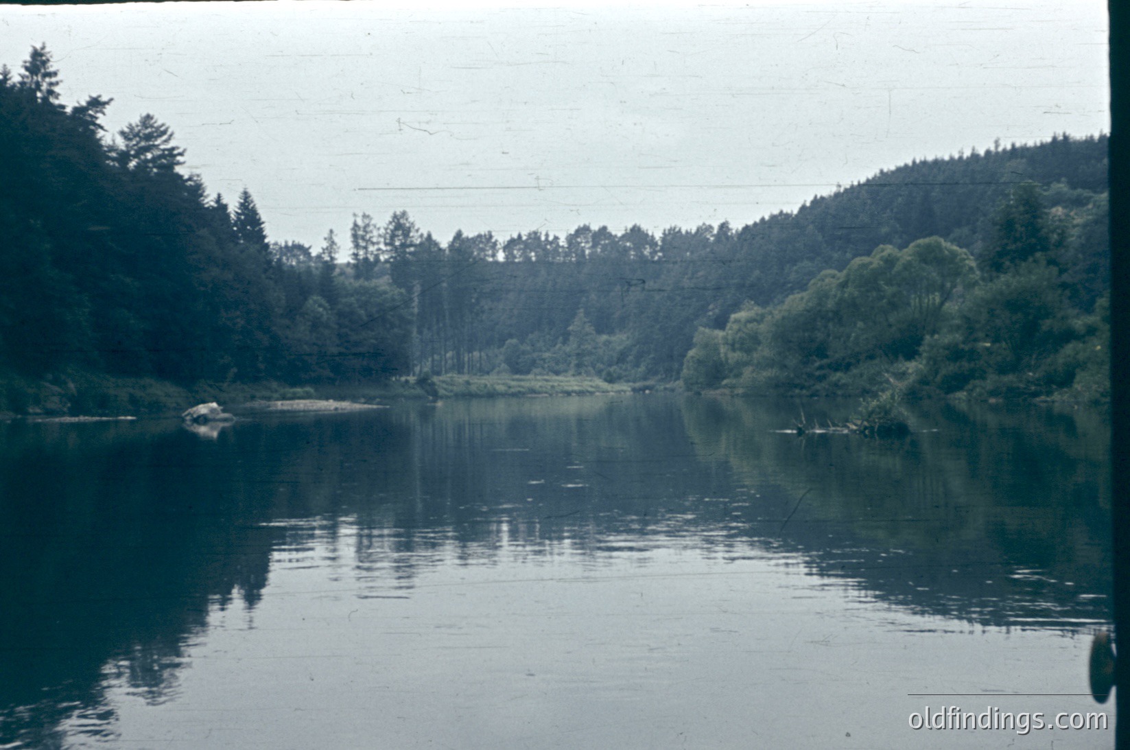 Calm lake scene with dense forest reflections, likely mid-20th century. Overcast skies and muted colors suggest vintage film grain. Dense coniferous trees frame both shores, with a lone structure visible in distance. Ideal for nature, travel, or historical stock imagery.